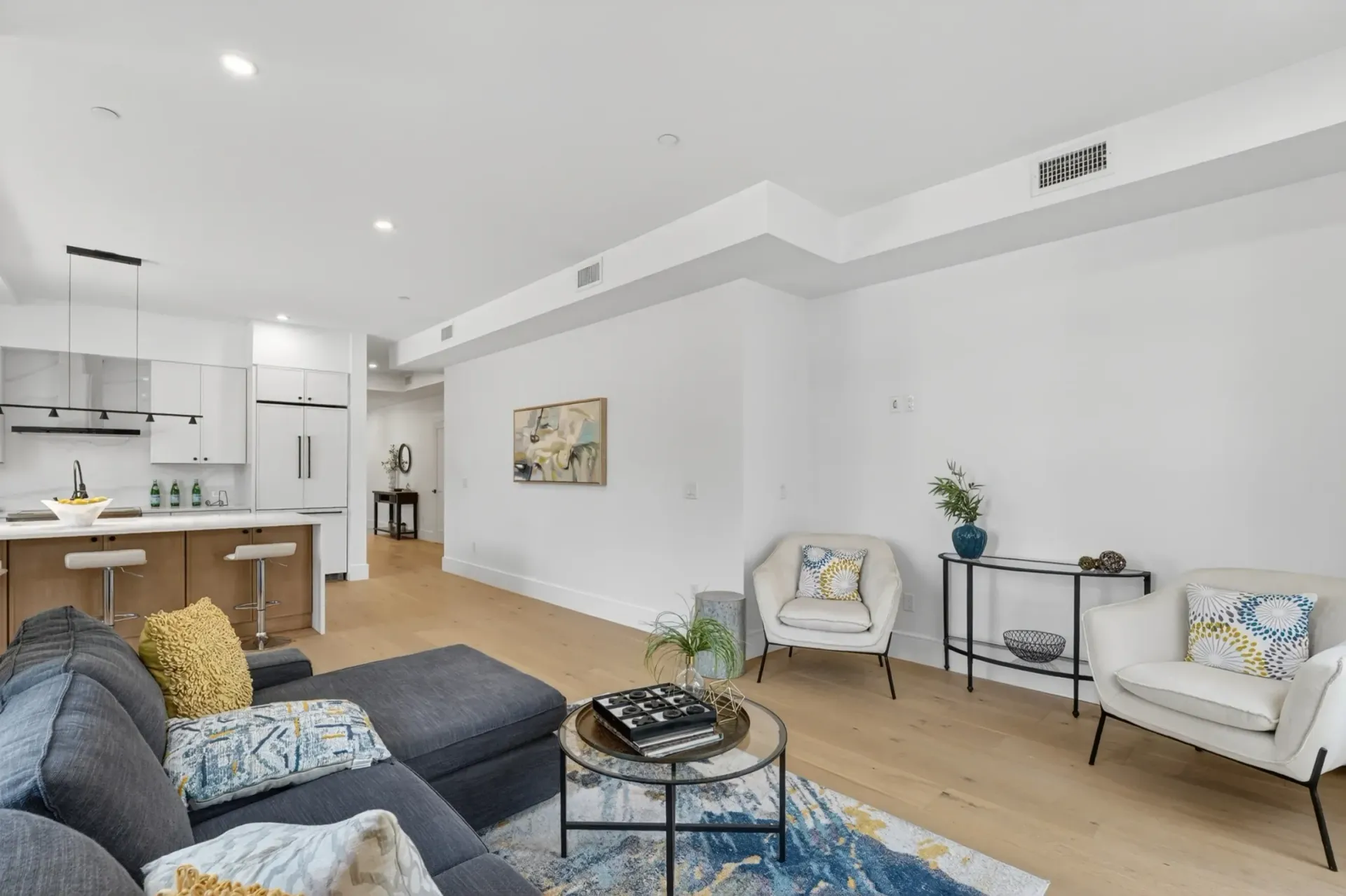 Bright open-concept living room with gray sofa, blue rug, white chairs, and kitchen bar stools