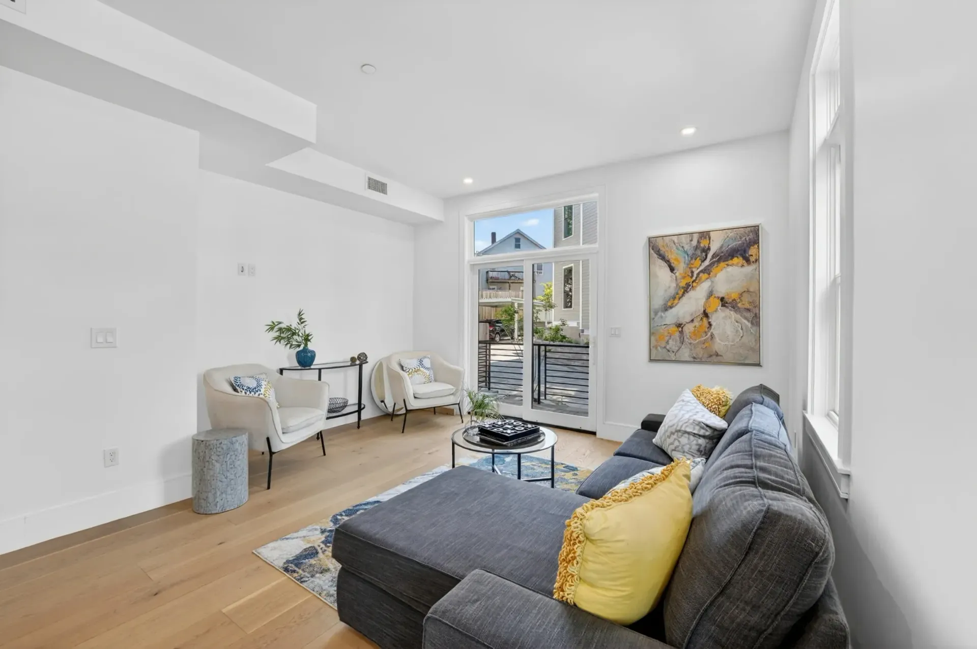 Bright modern living room with gray sofa, yellow pillows, white walls, and large windows.
