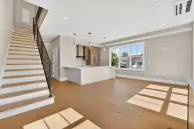 Bright empty living room with staircase, white kitchen, and sunlight through large windows on tan floor