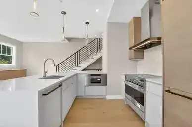Modern white kitchen with wood floors, island sink, stainless appliances, and staircase in the background