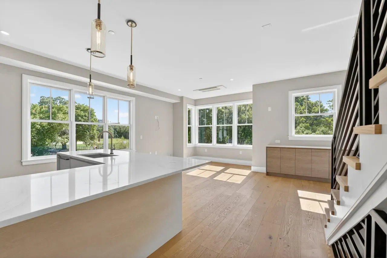 Bright open kitchen and living space with large windows, white island, wood floors, and staircase