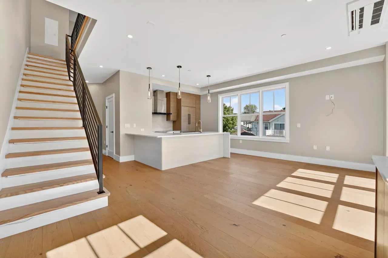 Bright empty modern living room with staircase, white kitchen island, wood floors, and sunlight through large windows