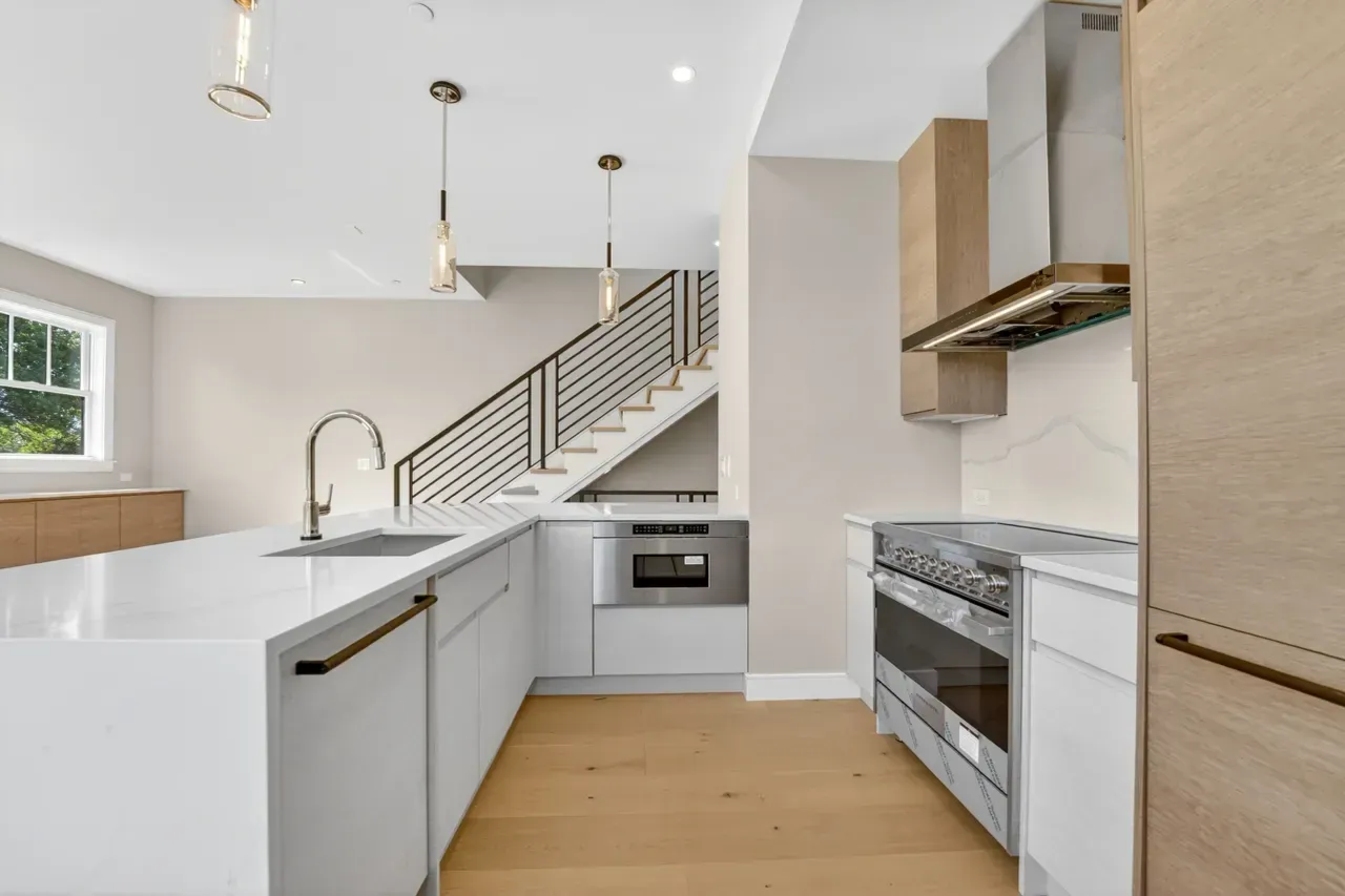 Modern white kitchen with wood floors, island sink, stainless appliances, and staircase in the background