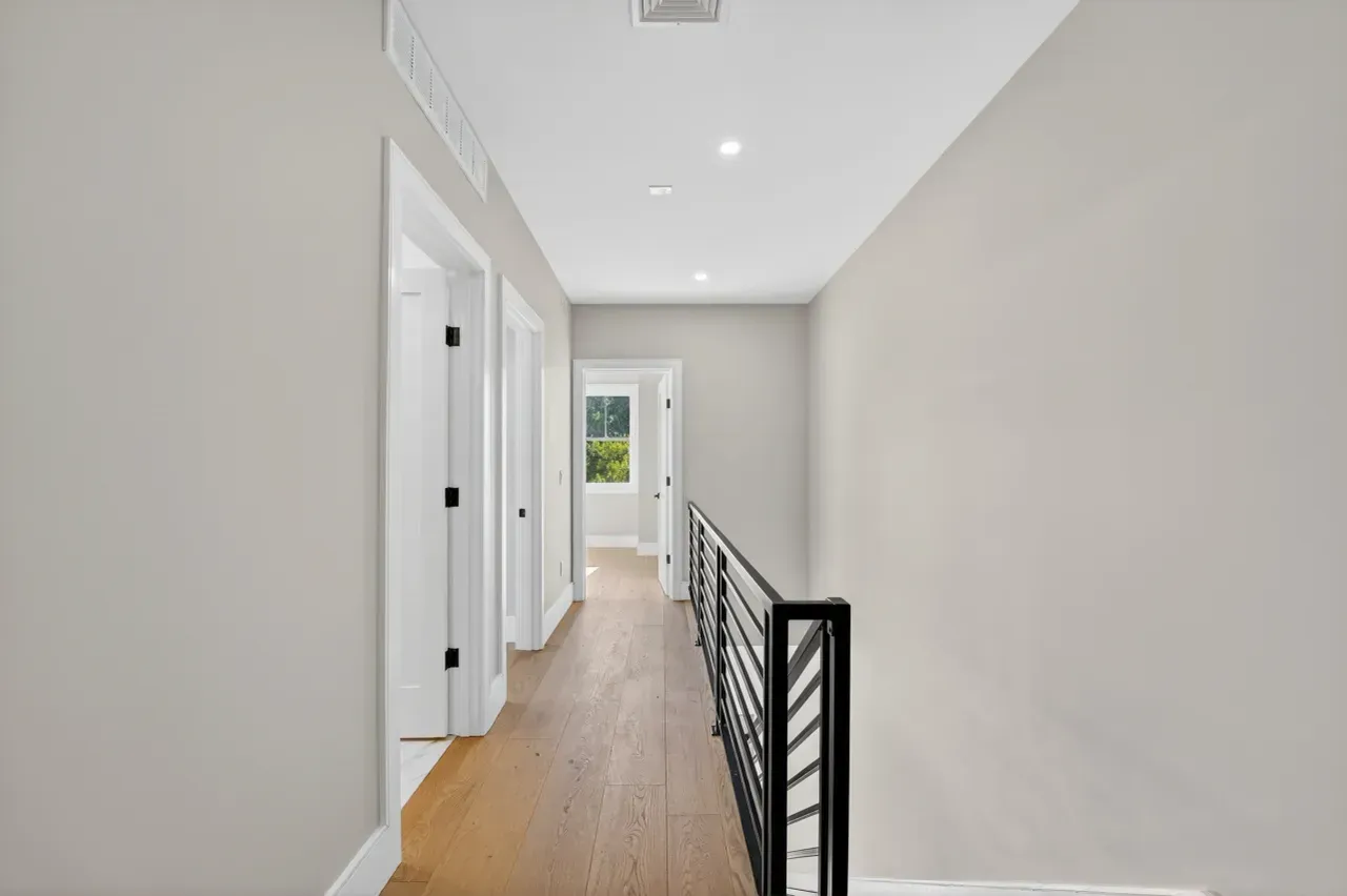 Bright hallway with white walls, hardwood floor, black railing, and a window at the far end