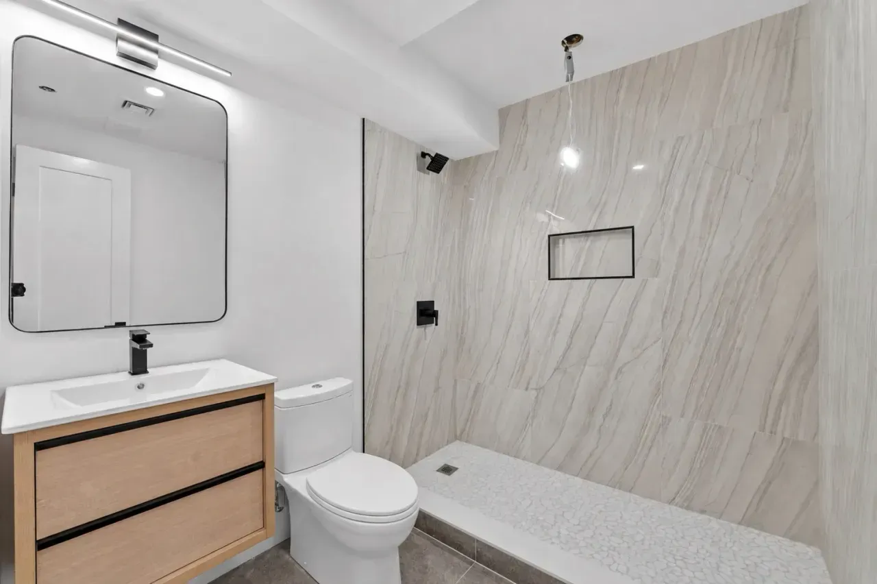 Modern bathroom with wood vanity, mirror, white toilet, and walk-in shower with large tile wall