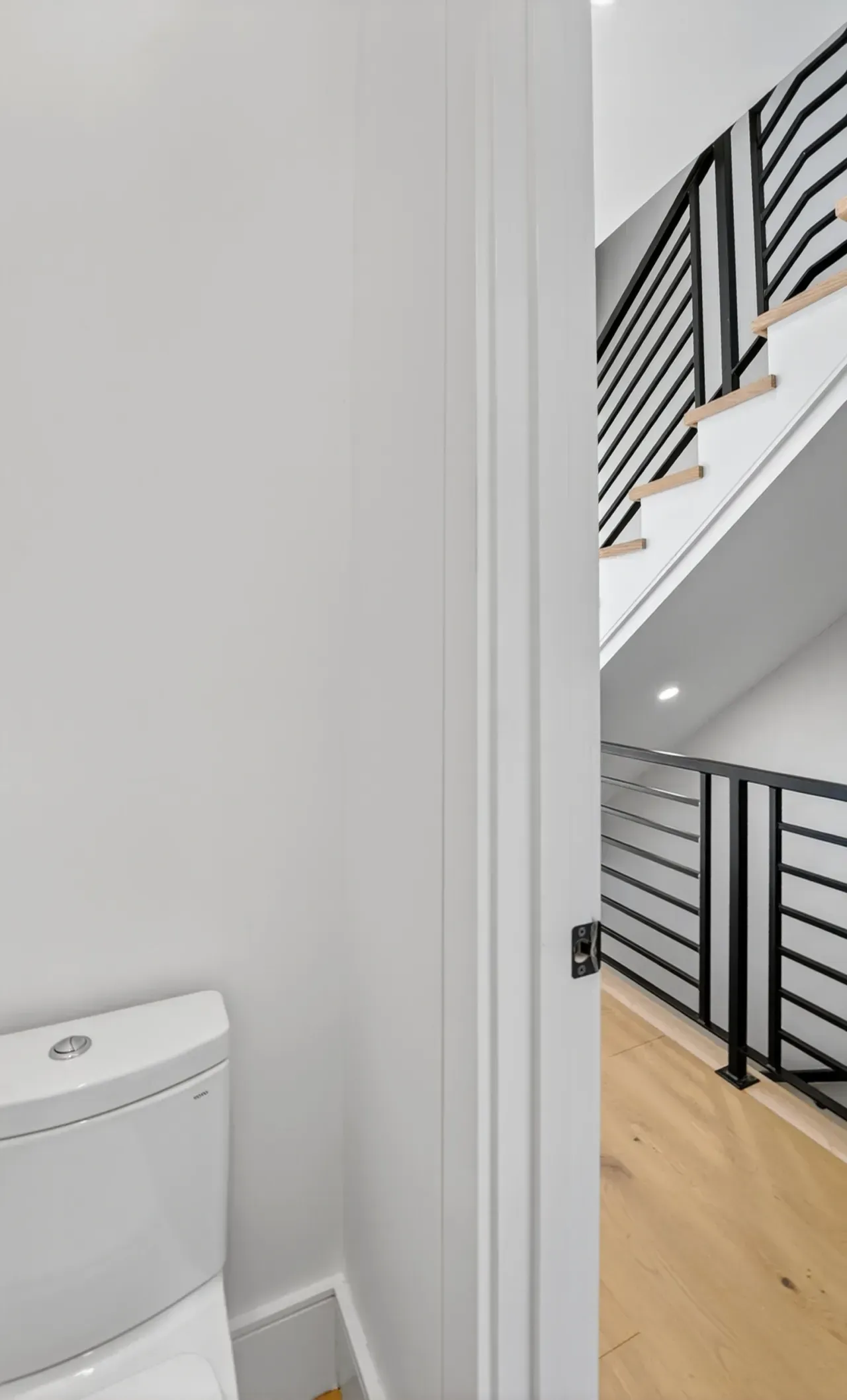 Small white bathroom beside a stairway with black railing and light wood floors
