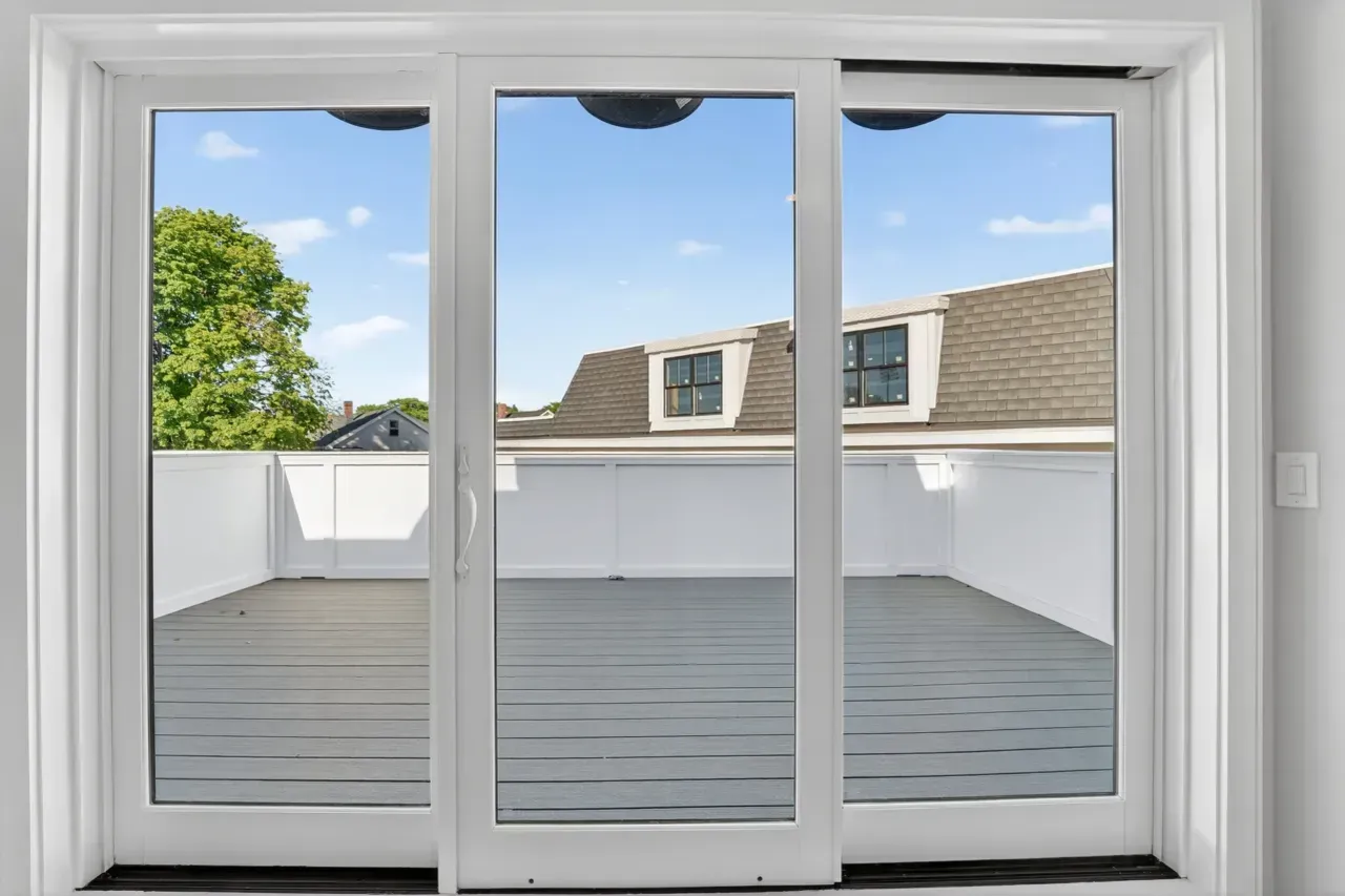 Sliding glass doors open to a rooftop deck with white railings and gray flooring under a blue sky