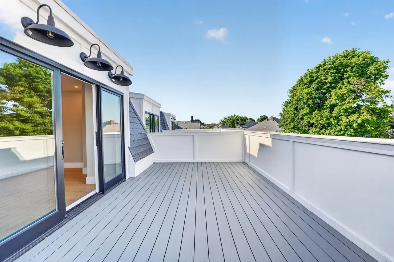 Spacious rooftop deck with gray flooring, white walls, glass doors, and trees beyond the railing