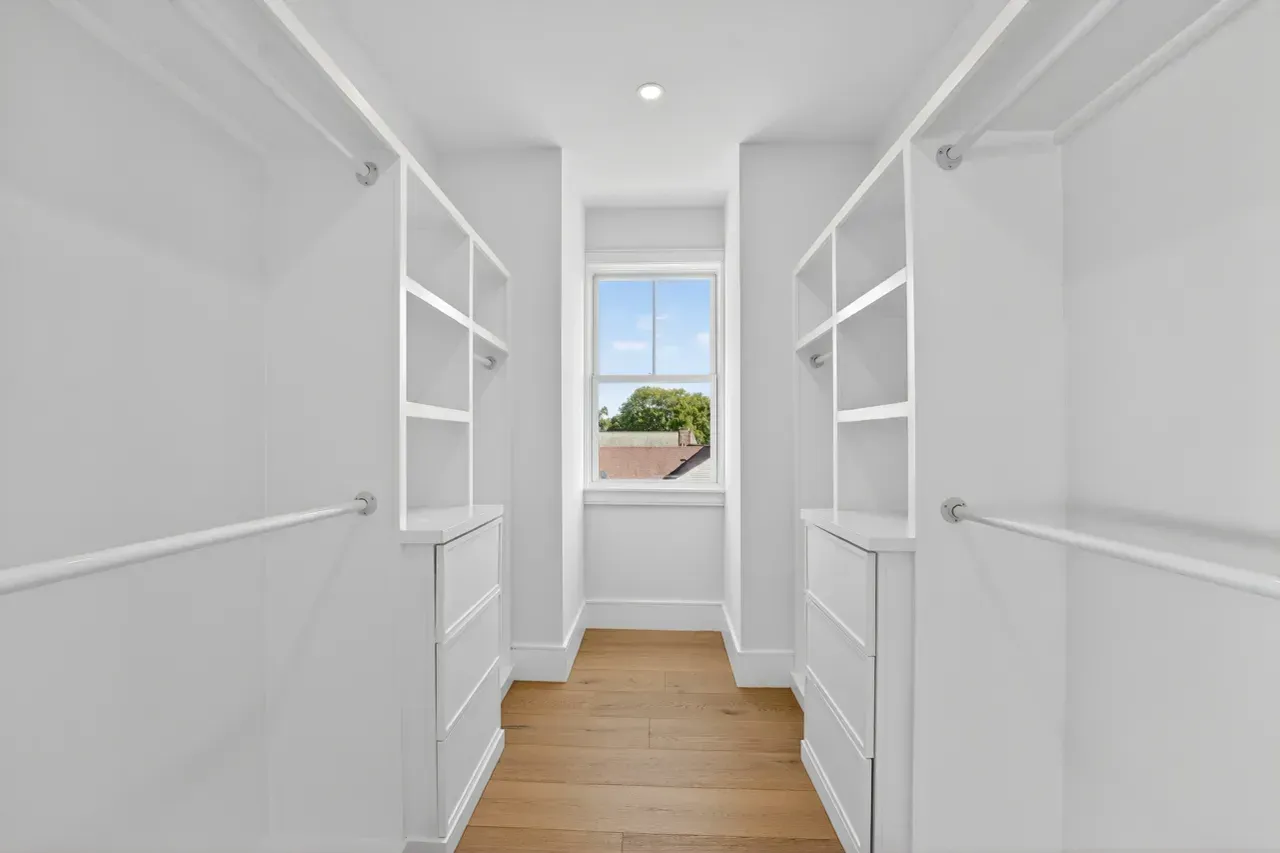 Bright white walk-in closet with built-in shelves, wood floor, and a window at the end