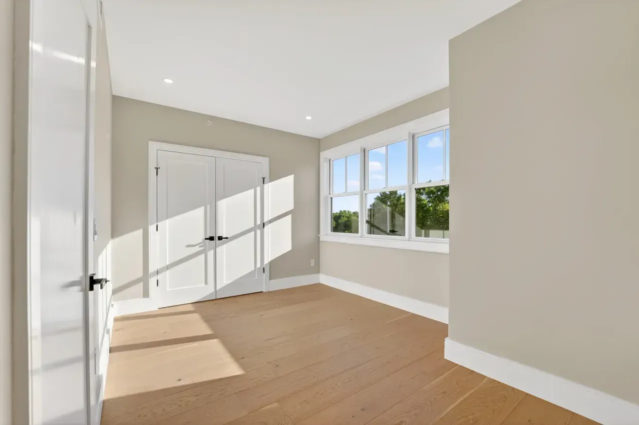 Empty sunlit bedroom with beige walls, wood floor, white closet doors, and a large window