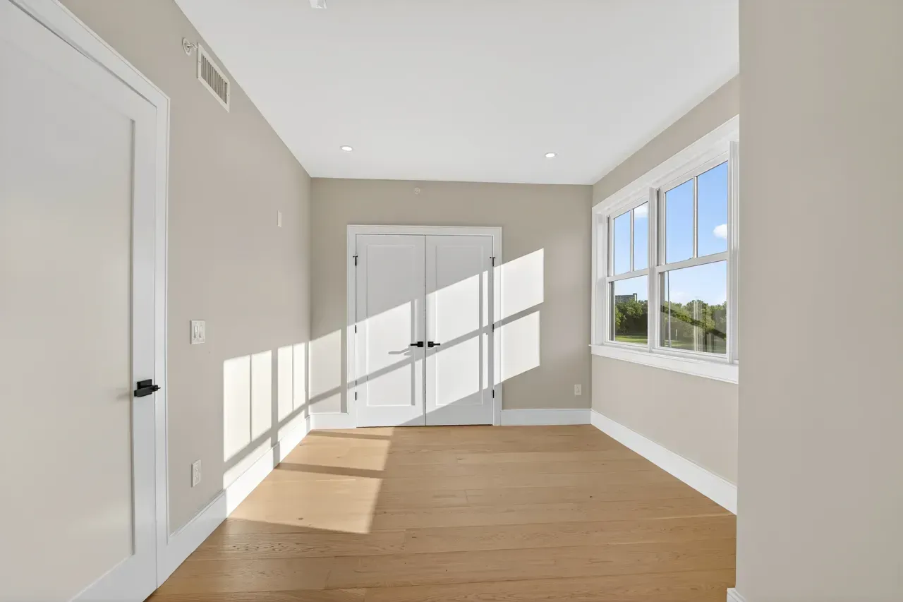 Bright empty room with hardwood floor, white doors, and sunlight streaming through large windows