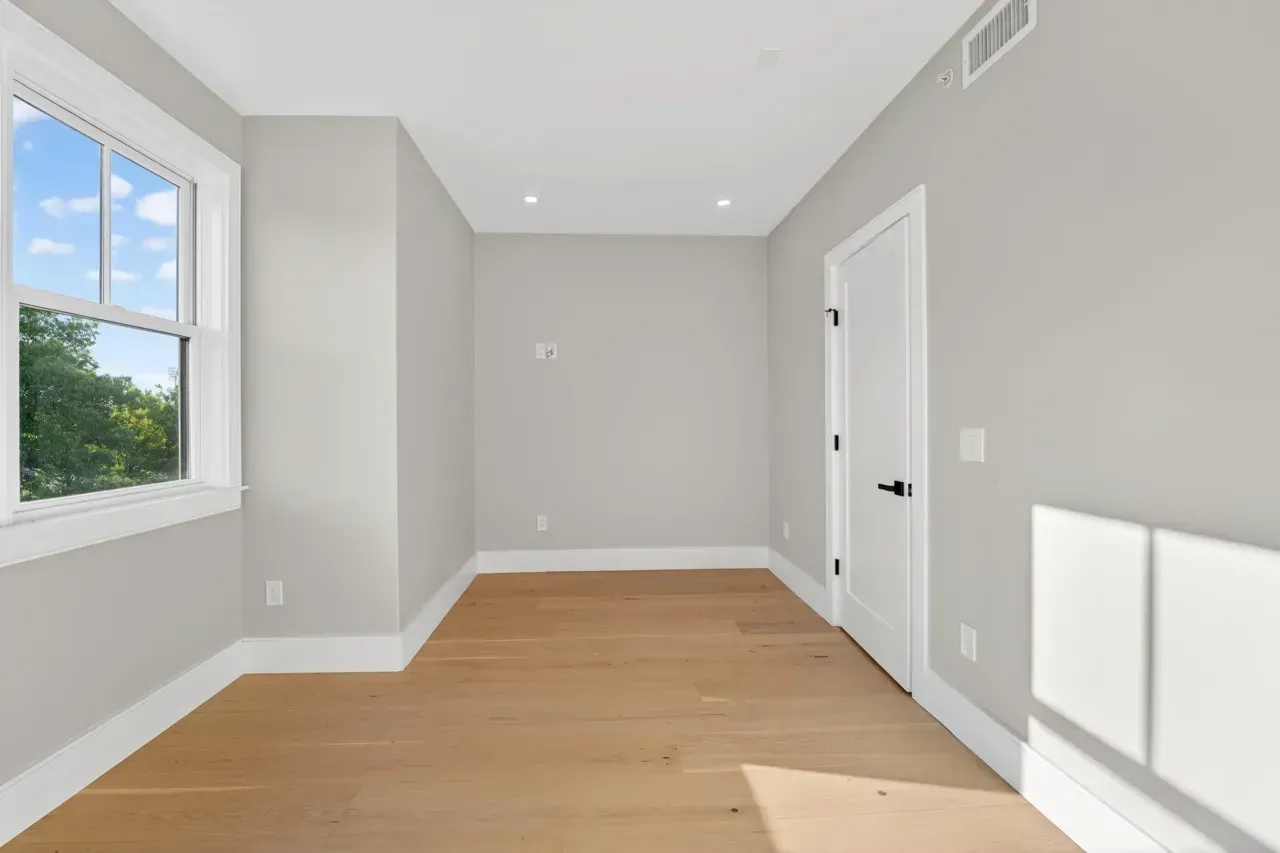Empty bright room with light wood floors, gray walls, a window, and sunlight on the right wall