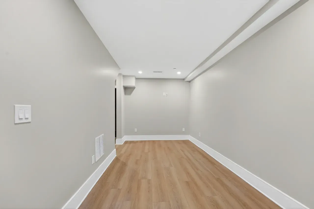 Empty narrow hallway with white walls, recessed ceiling lights, and light wood flooring