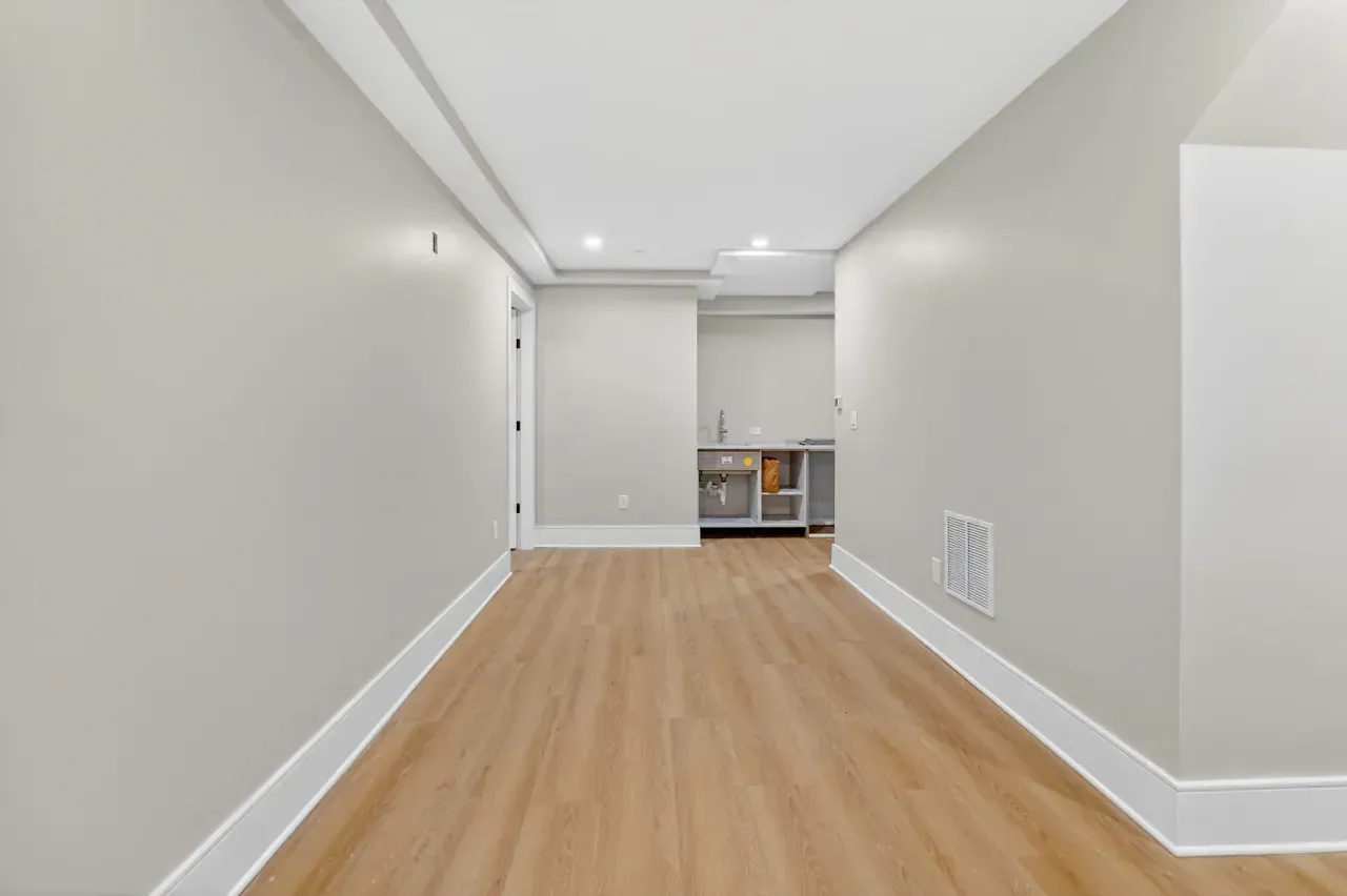 Empty narrow hallway with light wood floors and white walls, leading to a small room at the end