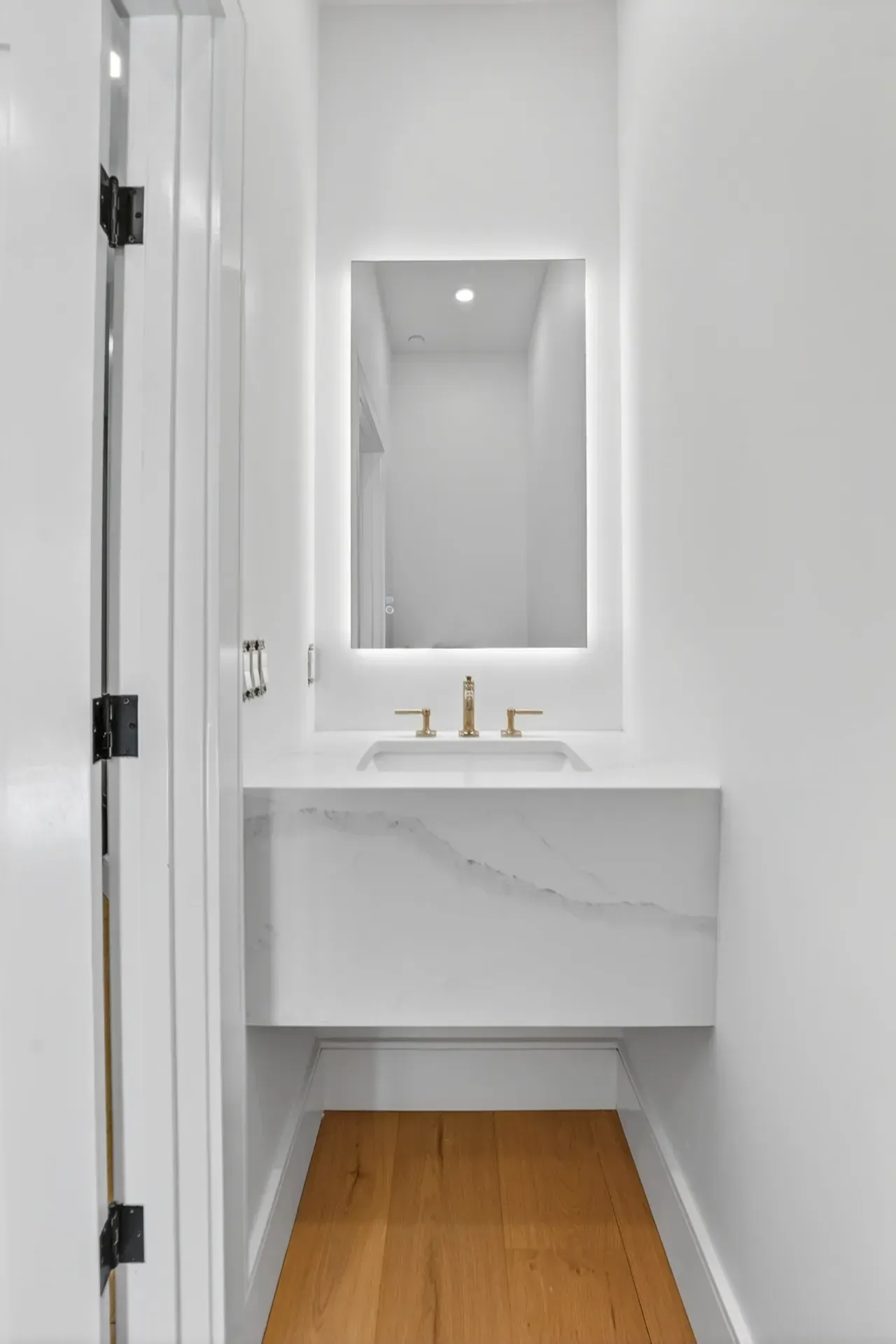 Narrow white bathroom vanity with gold faucet, mirror, and wood floor in a minimalist hallway