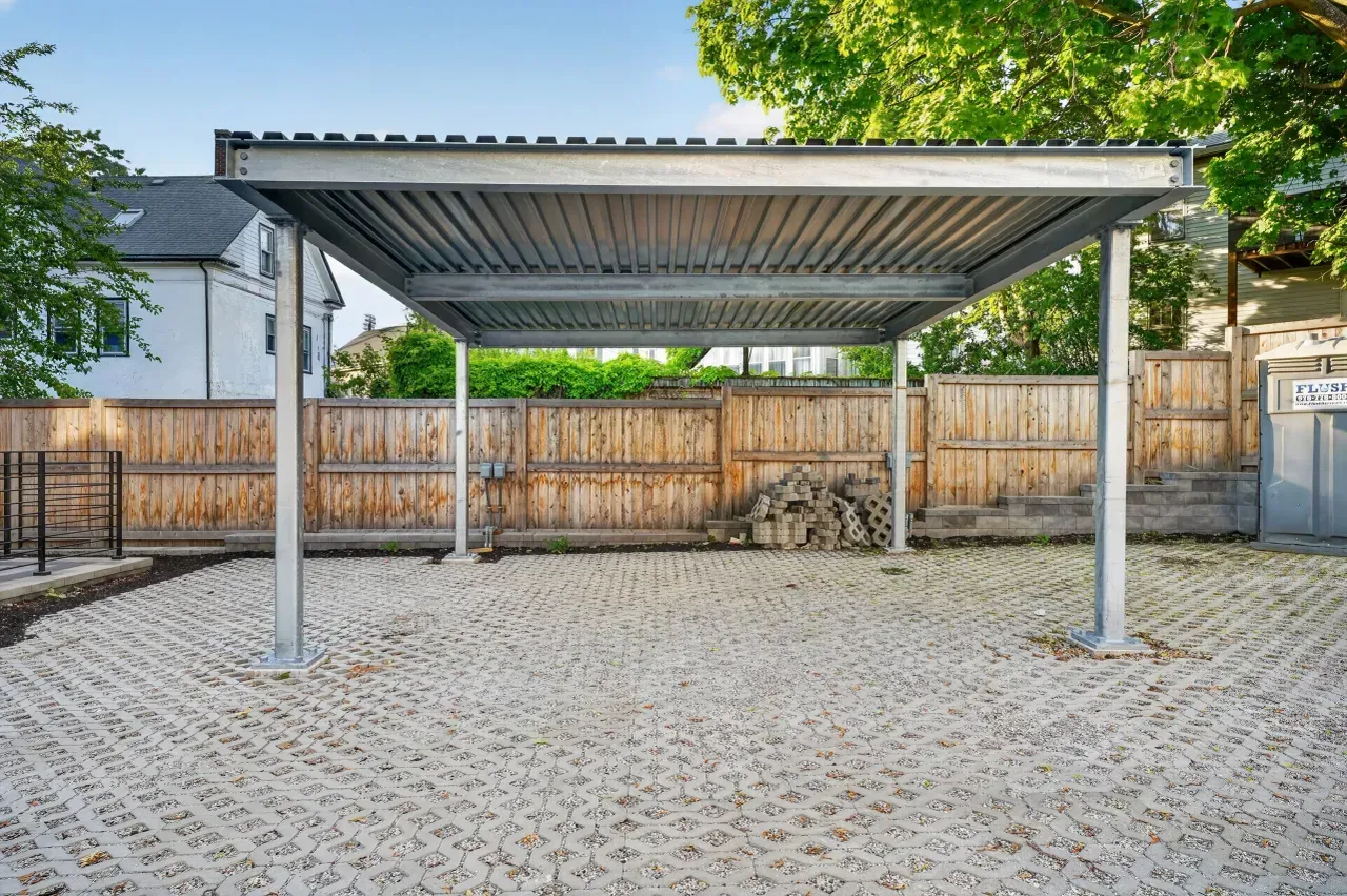 Covered carport with metal roof and four posts on a paved driveway, with a wooden fence behind it