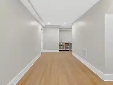 Empty modern room with light wood floor, white walls, and a small kitchen area at the far end