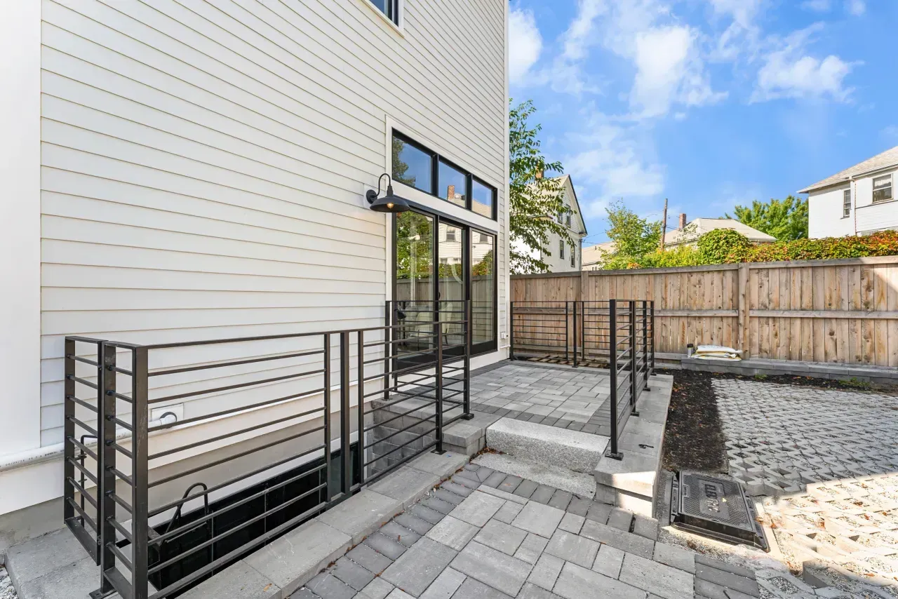 Modern house patio with paver walkway, black metal railing, and gravel yard beside a wood fence