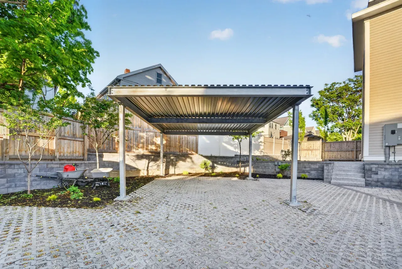 Empty metal carport in a paved courtyard beside a house and tree