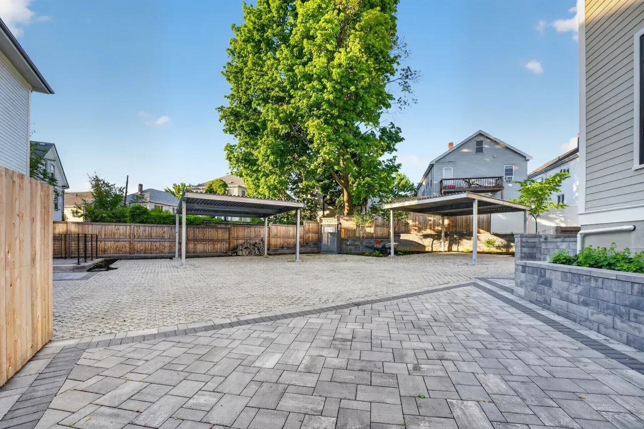 Paved backyard patio with a large tree, covered parking area, and neighboring houses in bright daylight