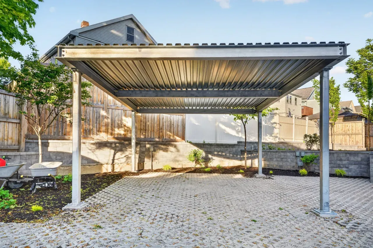 Covered carport with a metal roof and posts over a gravel driveway, beside a house and stone wall
