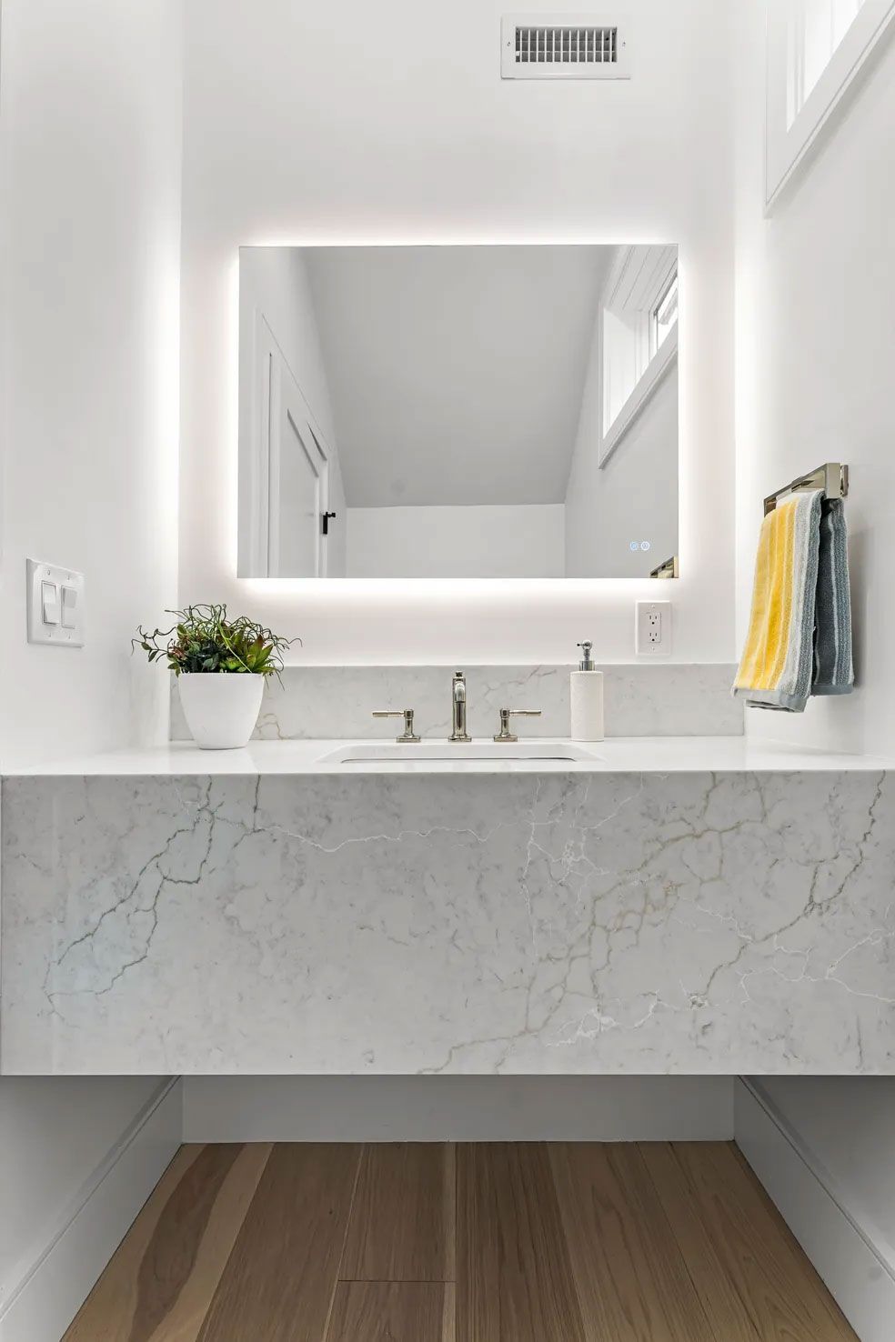 Modern white bathroom with floating marble vanity, lighted mirror, and wooden floor.