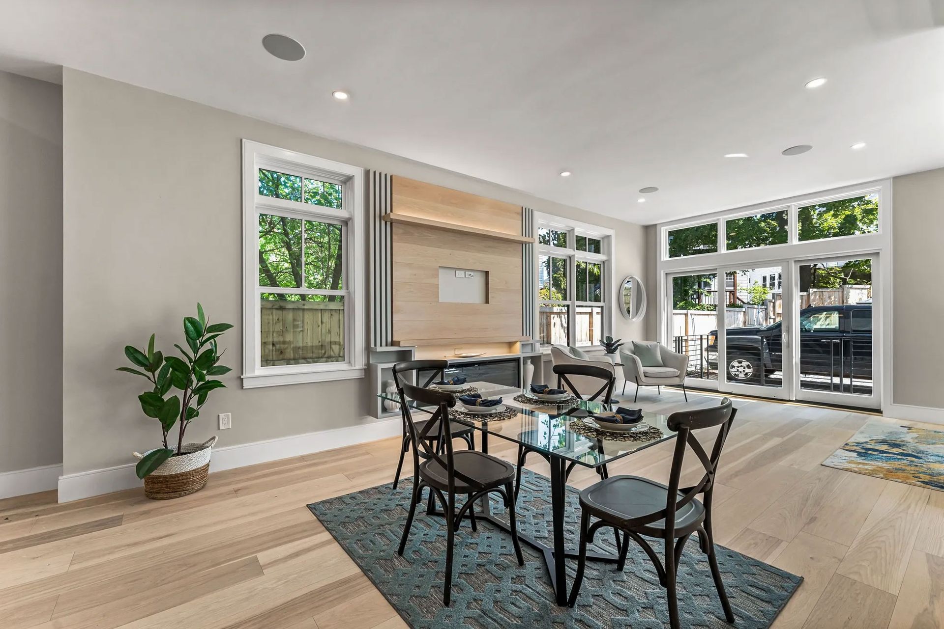 Dining room with wooden floor, table, and chairs. Large windows, potted plant, and light grey walls.