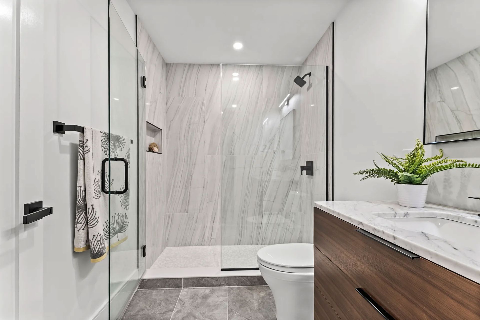 Modern bathroom with glass shower, white marble tile, wood vanity, and a potted fern.