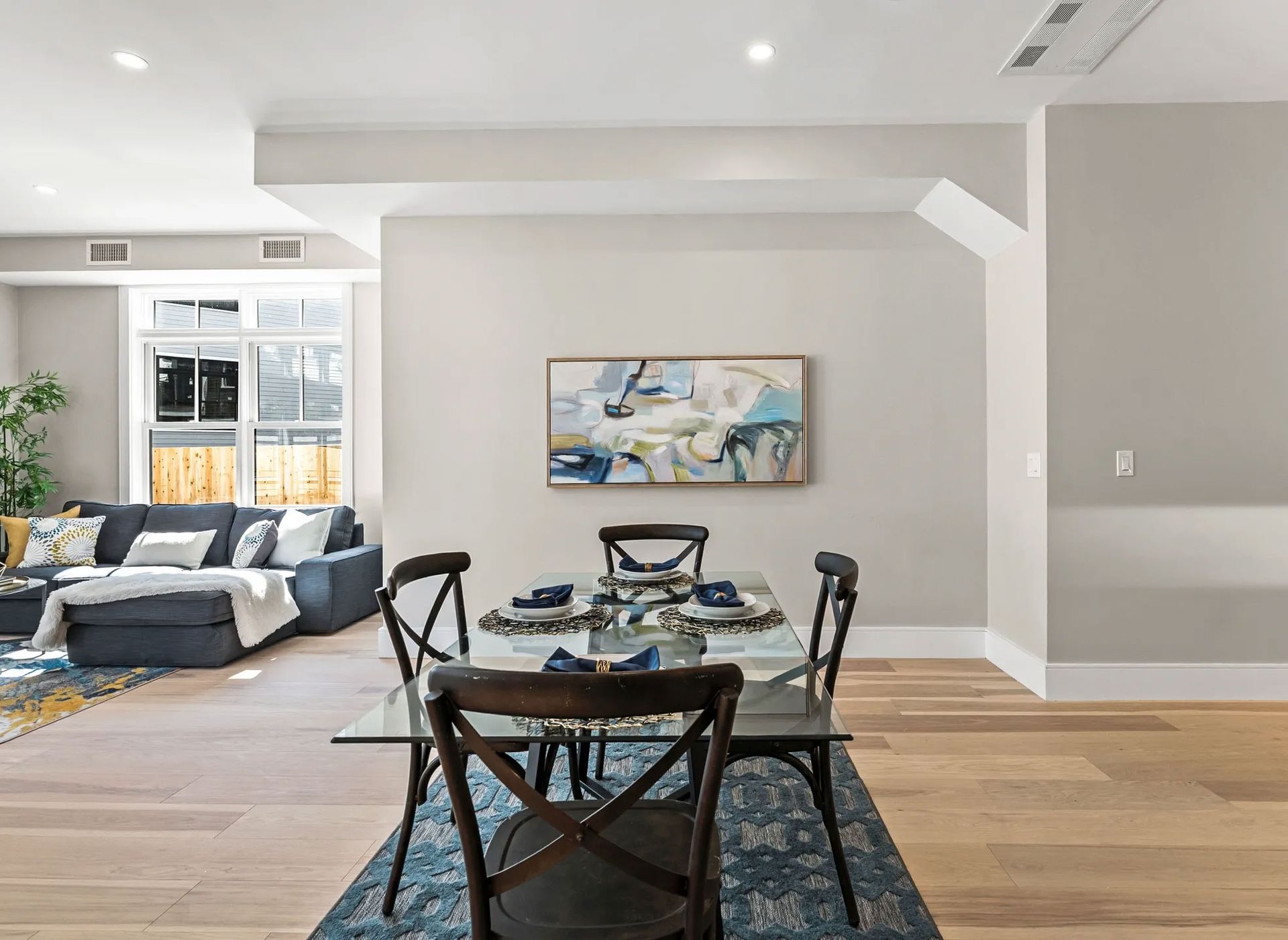 Dining room with glass table, four chairs, blue rug, and abstract art; living room visible.