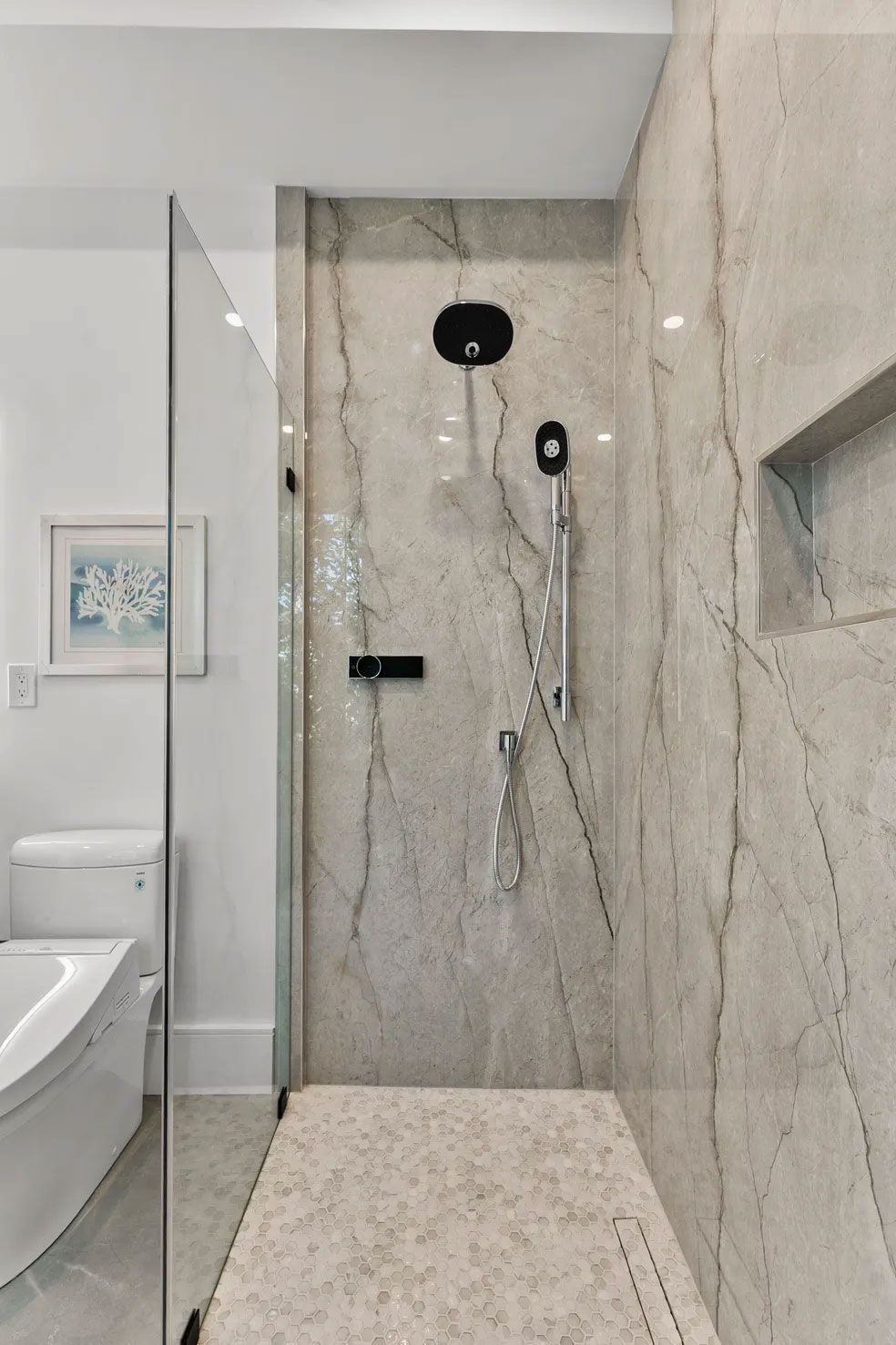 Modern marble-tiled shower with glass door, rain showerhead, handheld shower, and recessed shelf.