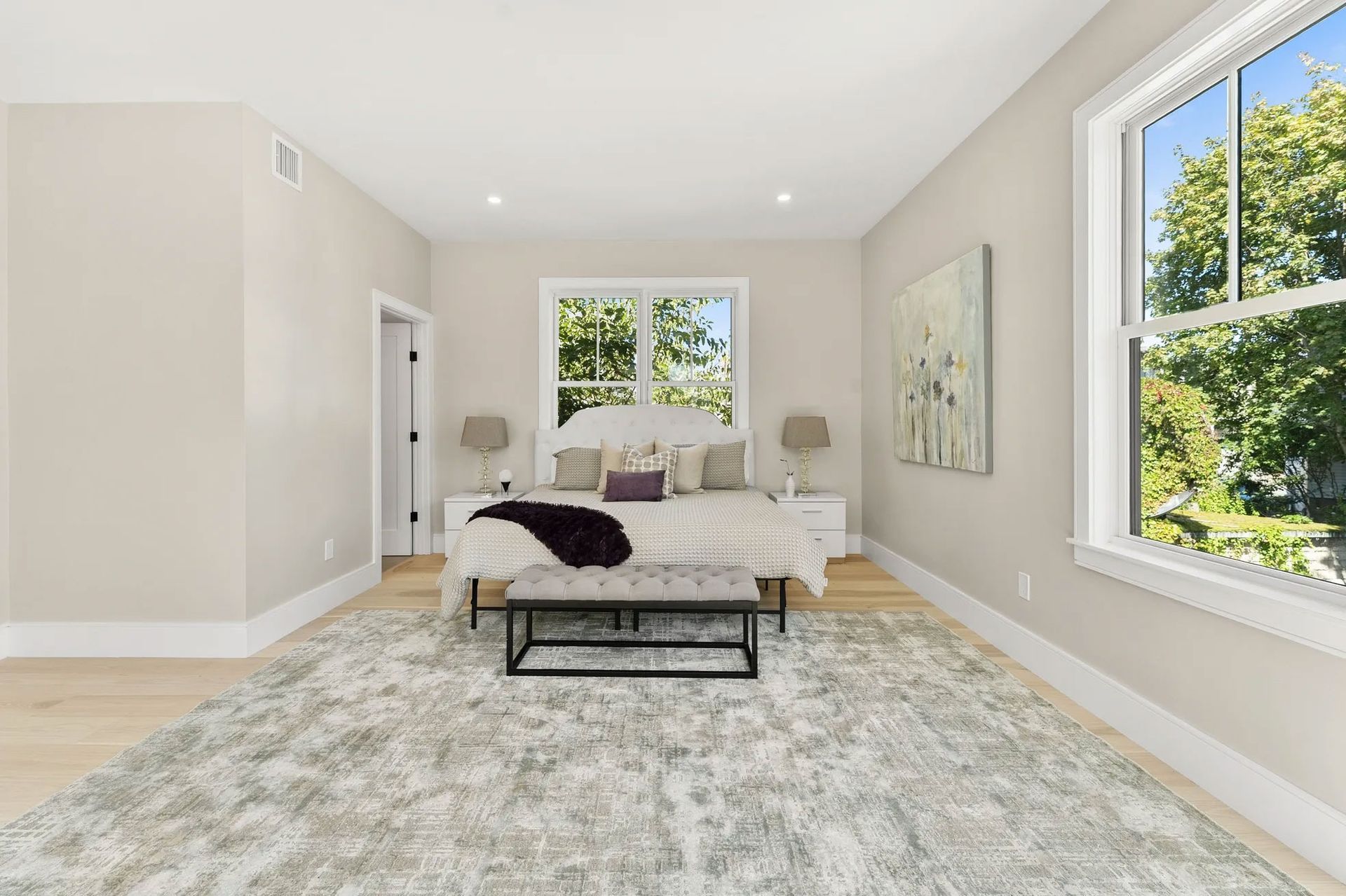 Bedroom with neutral walls, light wood floor, large rug, bed, two windows, and bench.