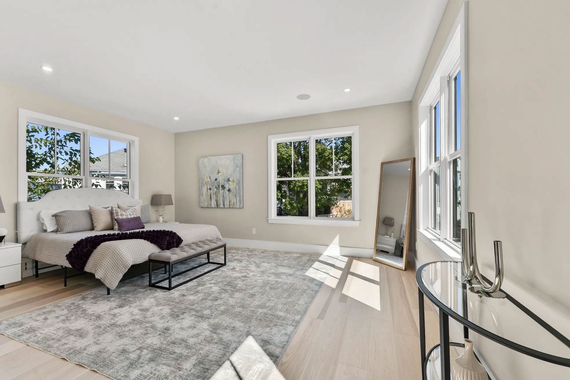Bright bedroom with large windows, neutral walls, plush rug, bed with purple accents, and a full-length mirror.