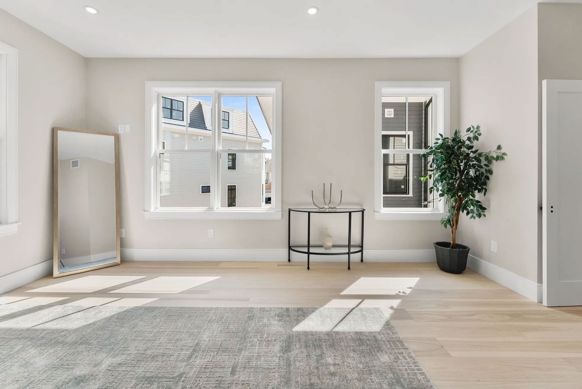 Empty room with large mirror, windows, rug, and plant. Light wood floors, white walls.