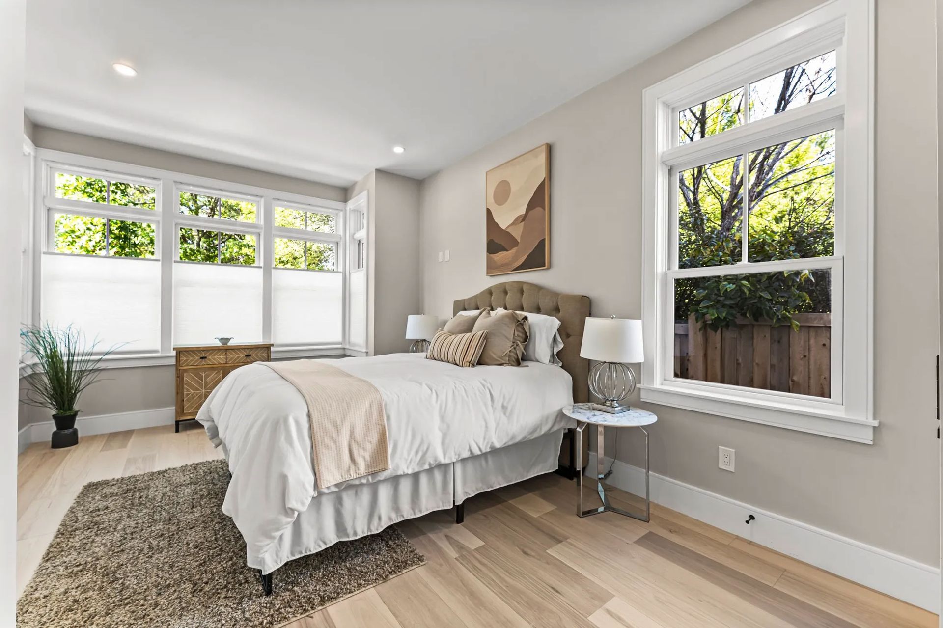 Bedroom with bed, windows, rug, and light-colored wood floor; neutral tones.