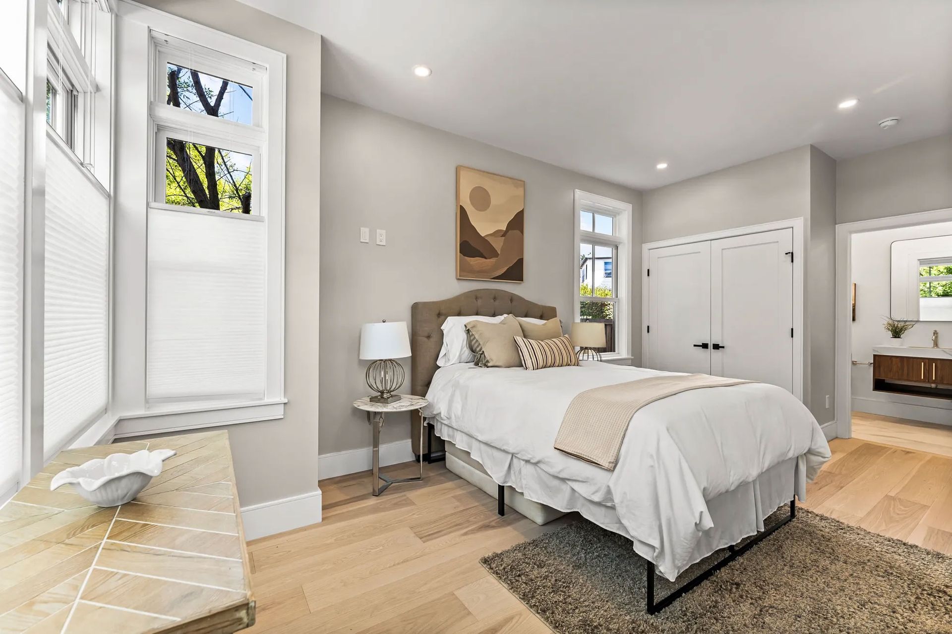 Bedroom with a bed, small side table, artwork, two windows, and closet. Pale gray walls and light wood floors.