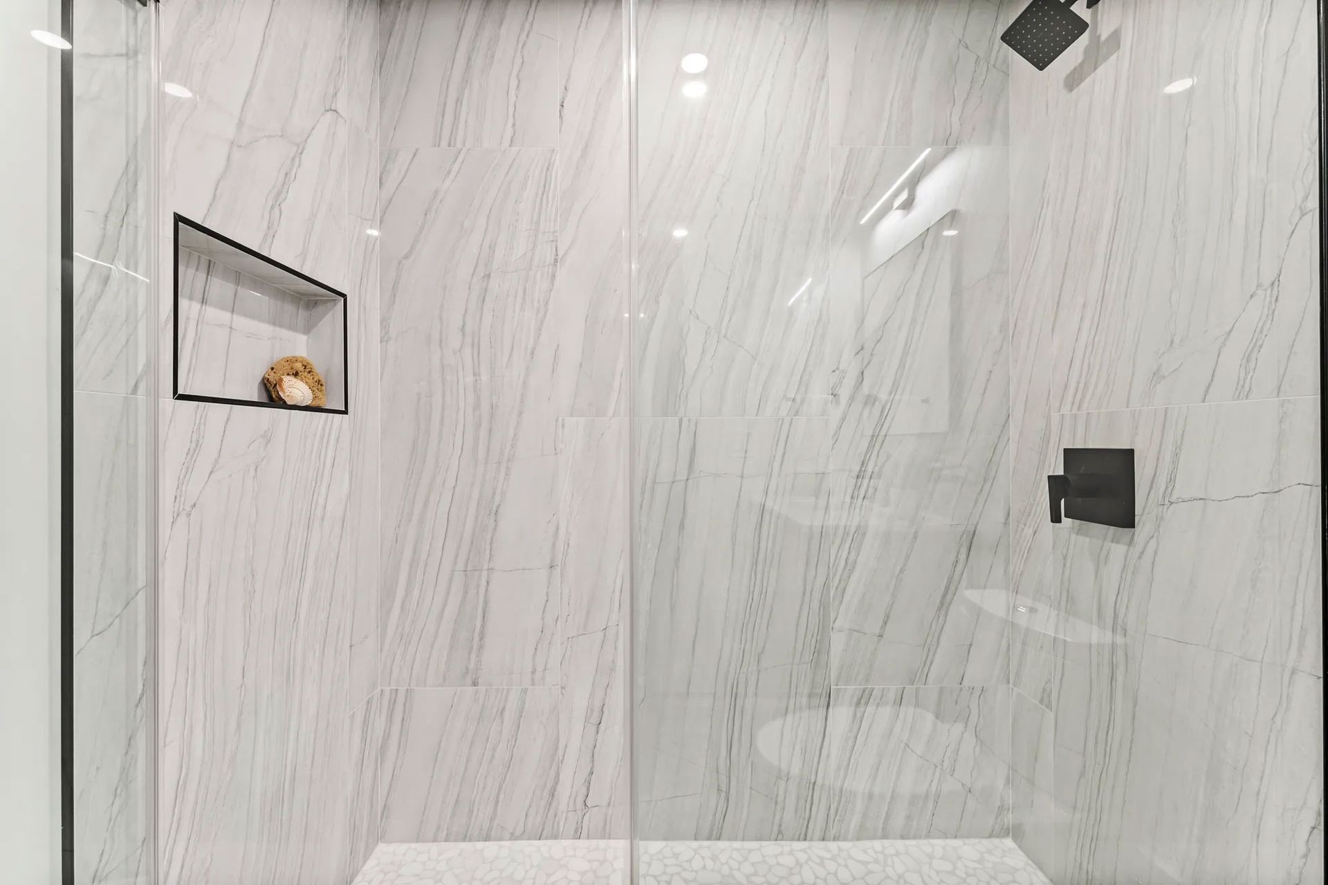 Modern shower with white marble walls, glass door, black fixtures, and a recessed shelf.