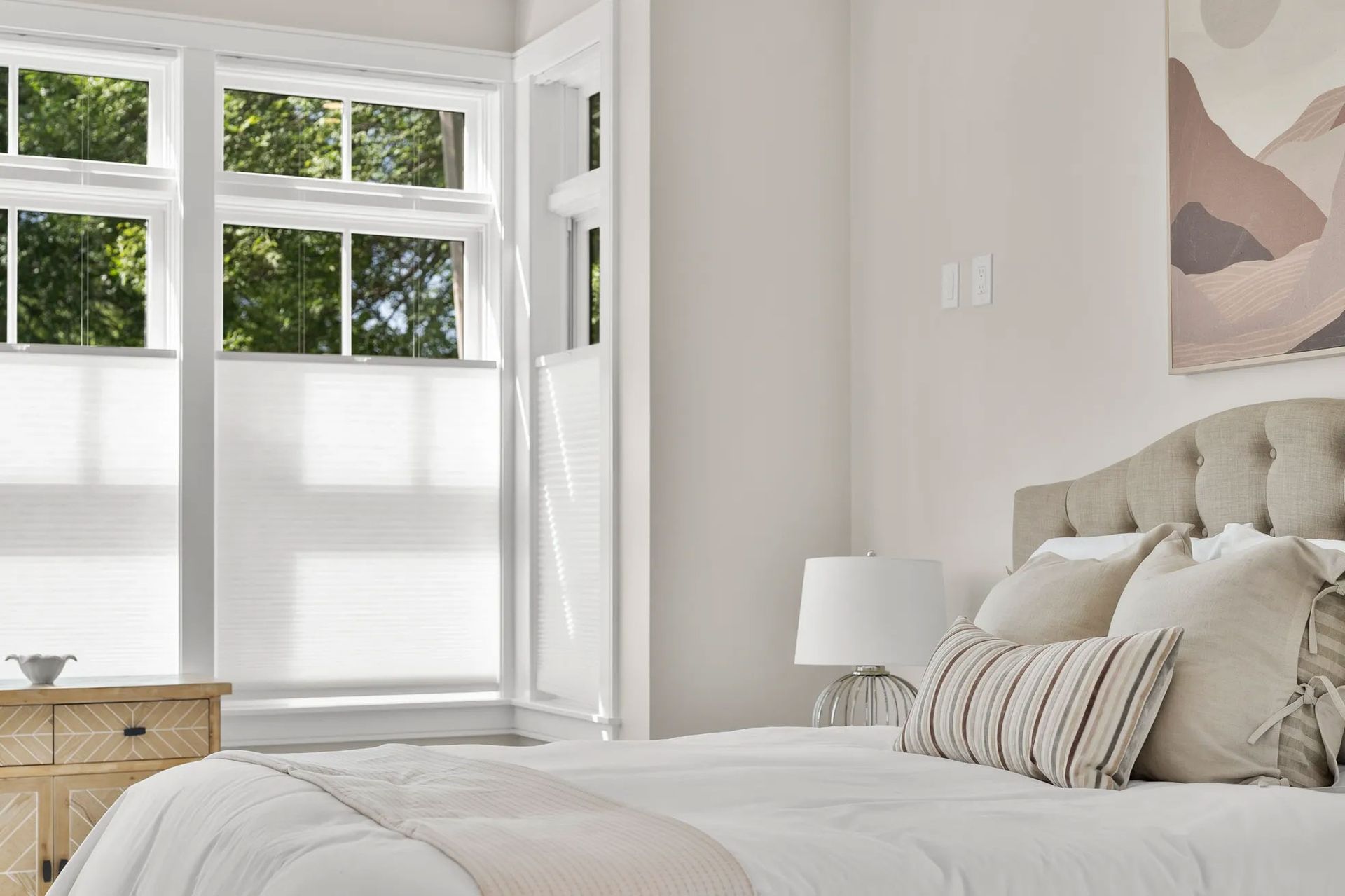 Bedroom with white bed, beige headboard, window with blinds, and artwork.