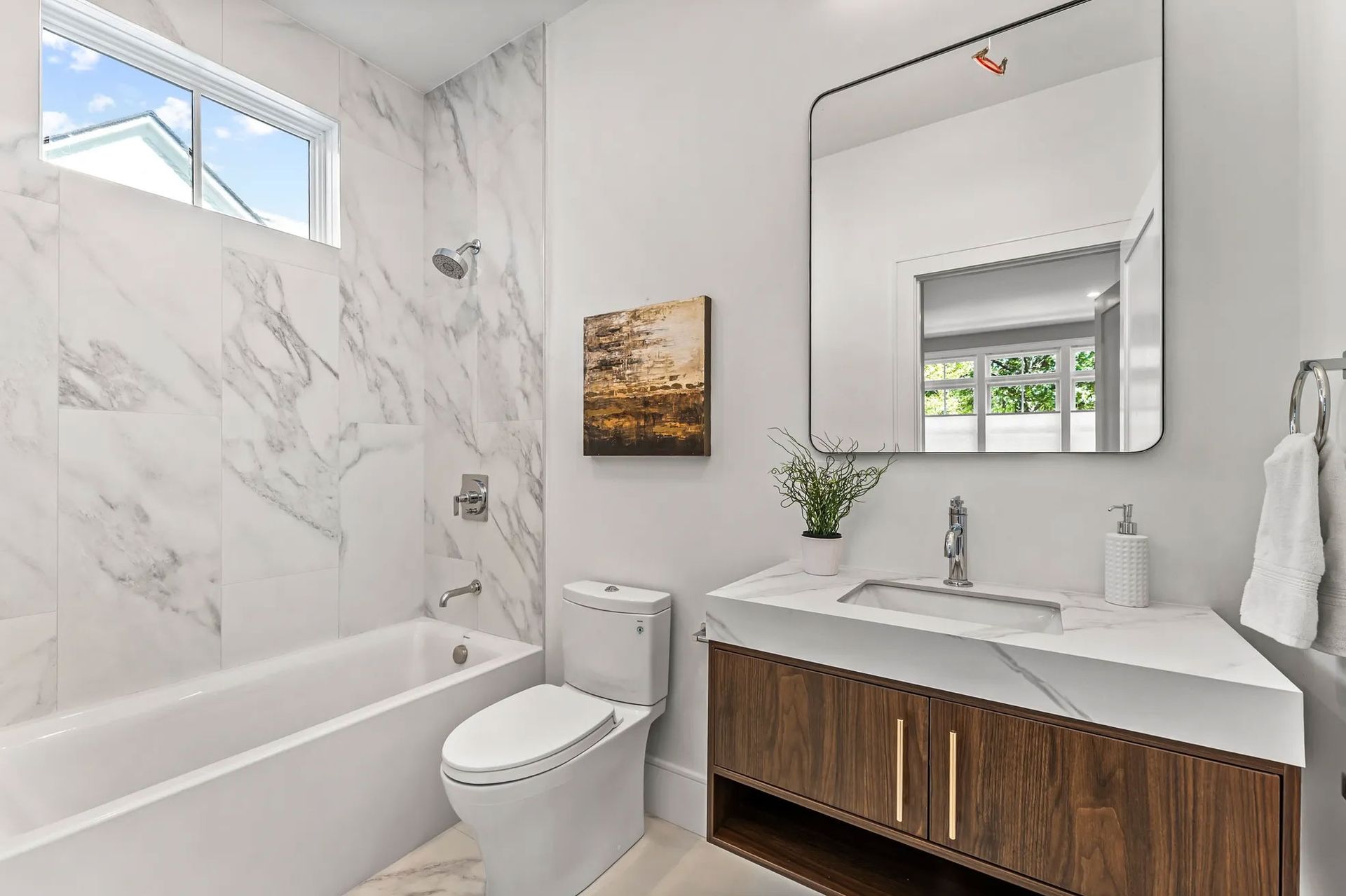 Modern bathroom with marble-tiled walls, a bathtub, toilet, vanity, and a large mirror.