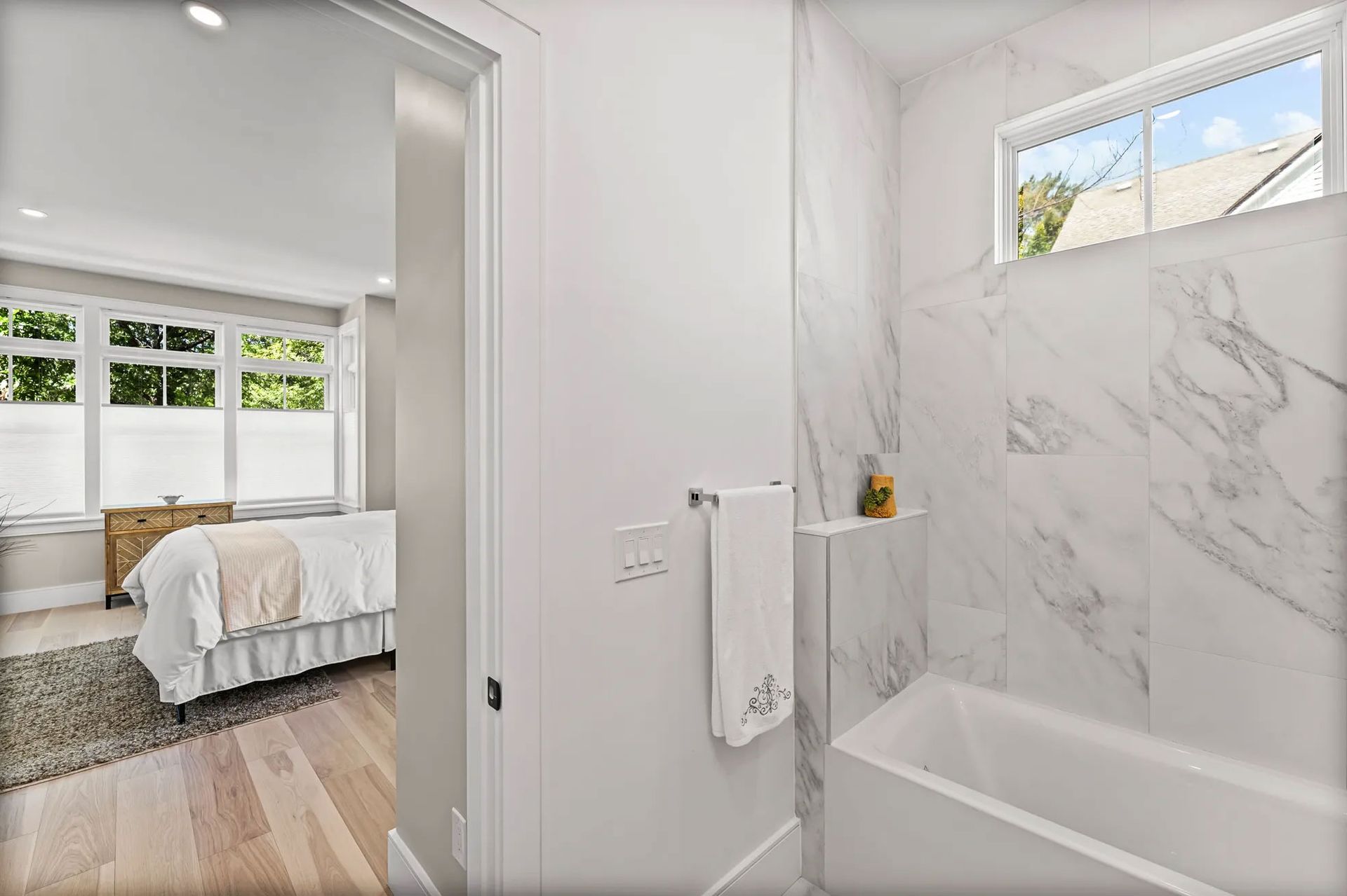 Bathroom with marble tile, a bathtub, and a window. Bedroom visible through the doorway with a bed.