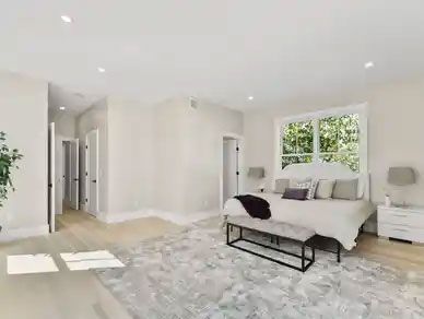 Spacious, bright bedroom with a bed, bench, rug, and large window; several doors lead to other rooms.