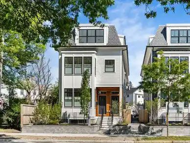 White two-story house with black trim, gray roof, and a small front yard, surrounded by trees.