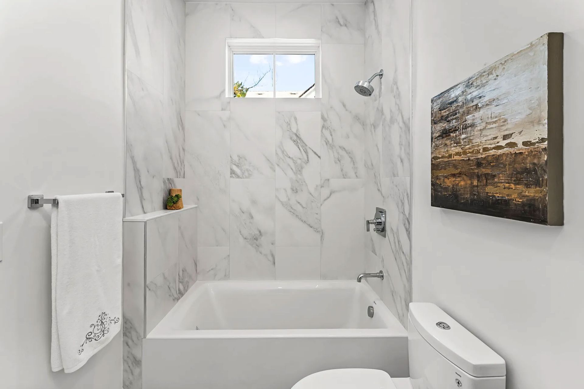 Bathroom with marble-like tile, white tub, toilet, and an abstract painting. A towel hangs on a chrome rack.