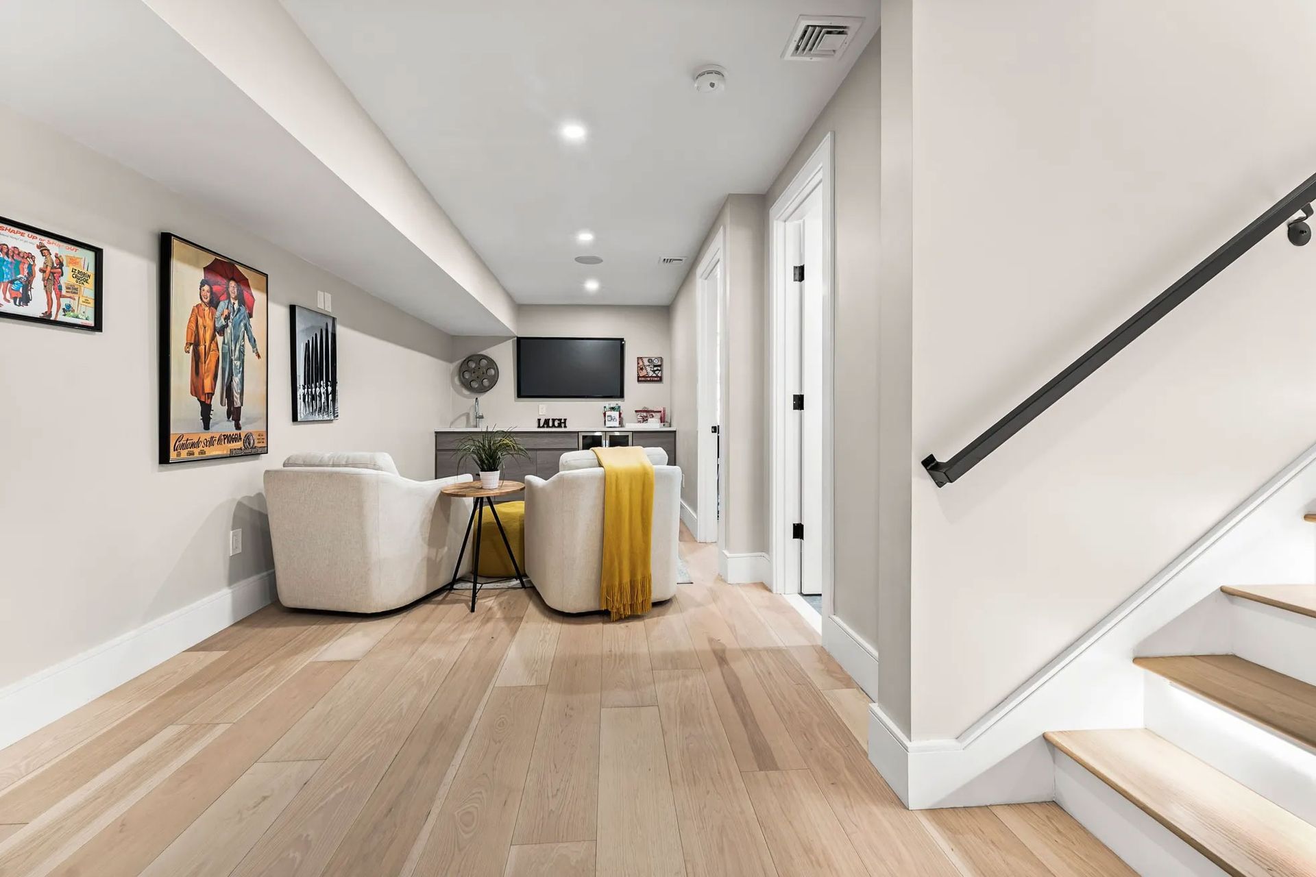Basement living room with light wood floors, seating area, and stairs leading up.