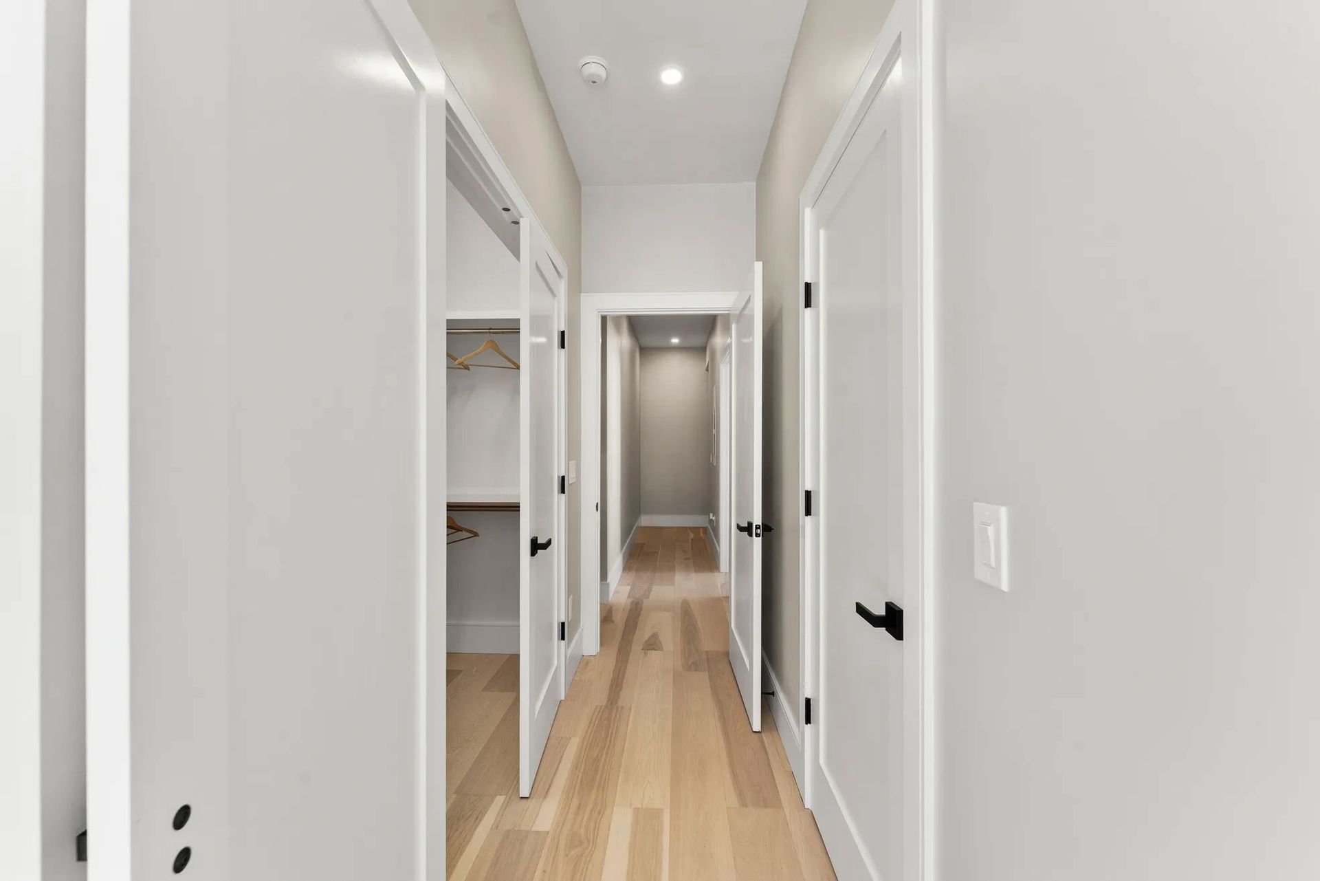 Narrow hallway with light wood floor, white walls and doors. One door open to a closet.