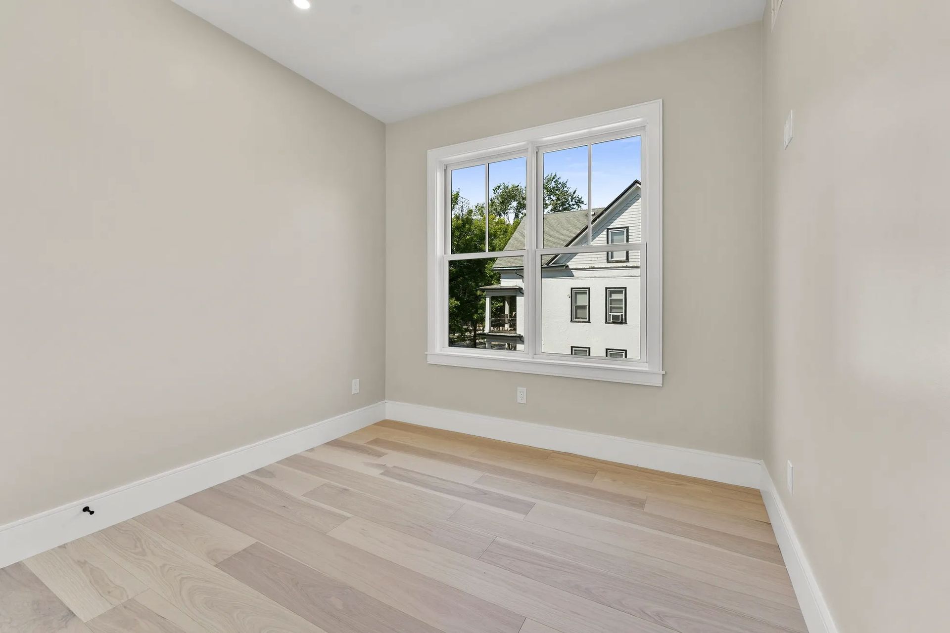 Empty room with light-colored wood flooring, white trim, and a window overlooking a tree-lined street.