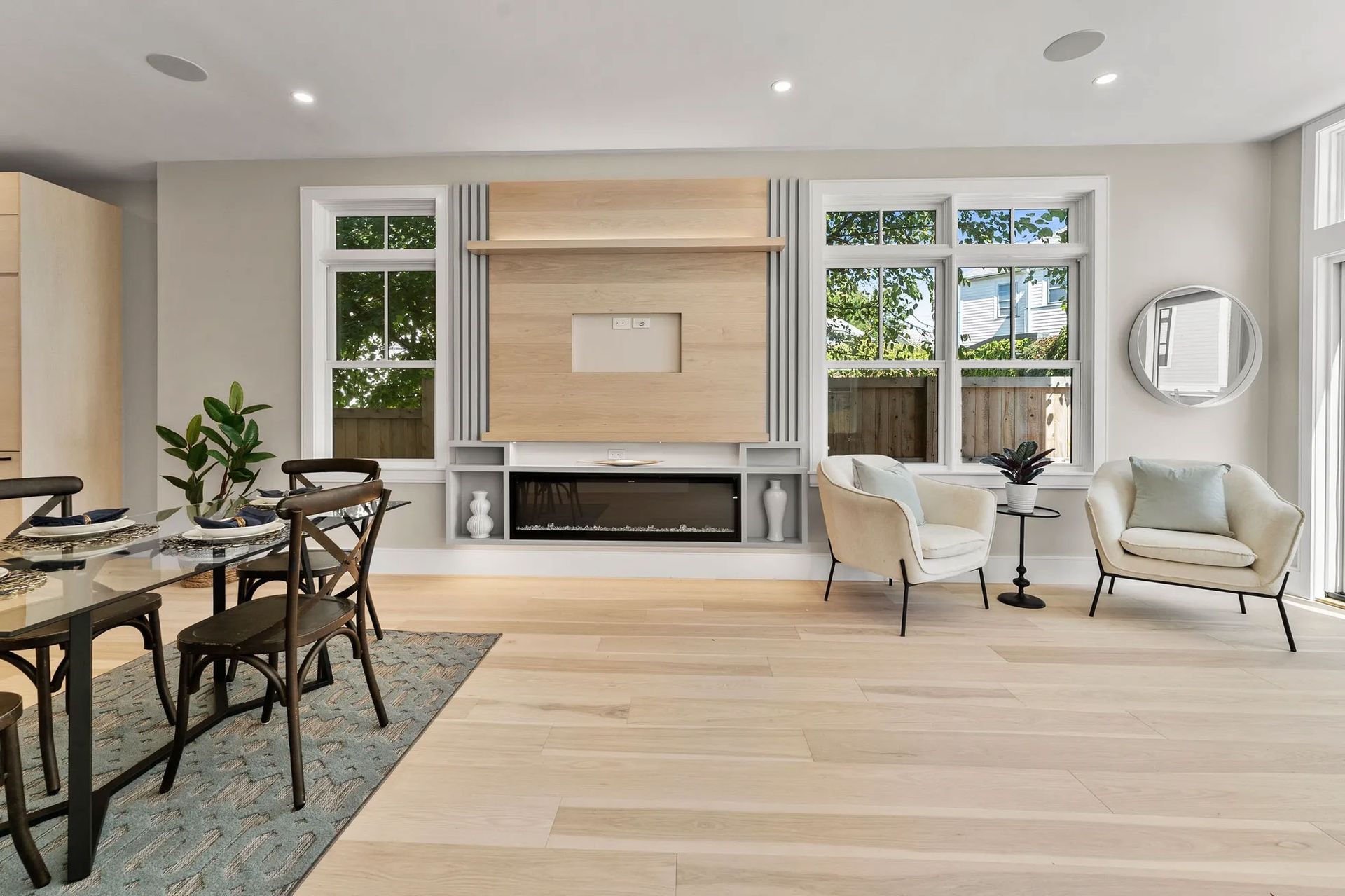 Bright living room with fireplace, windows, and light wood flooring. Dining table and chairs on the left.