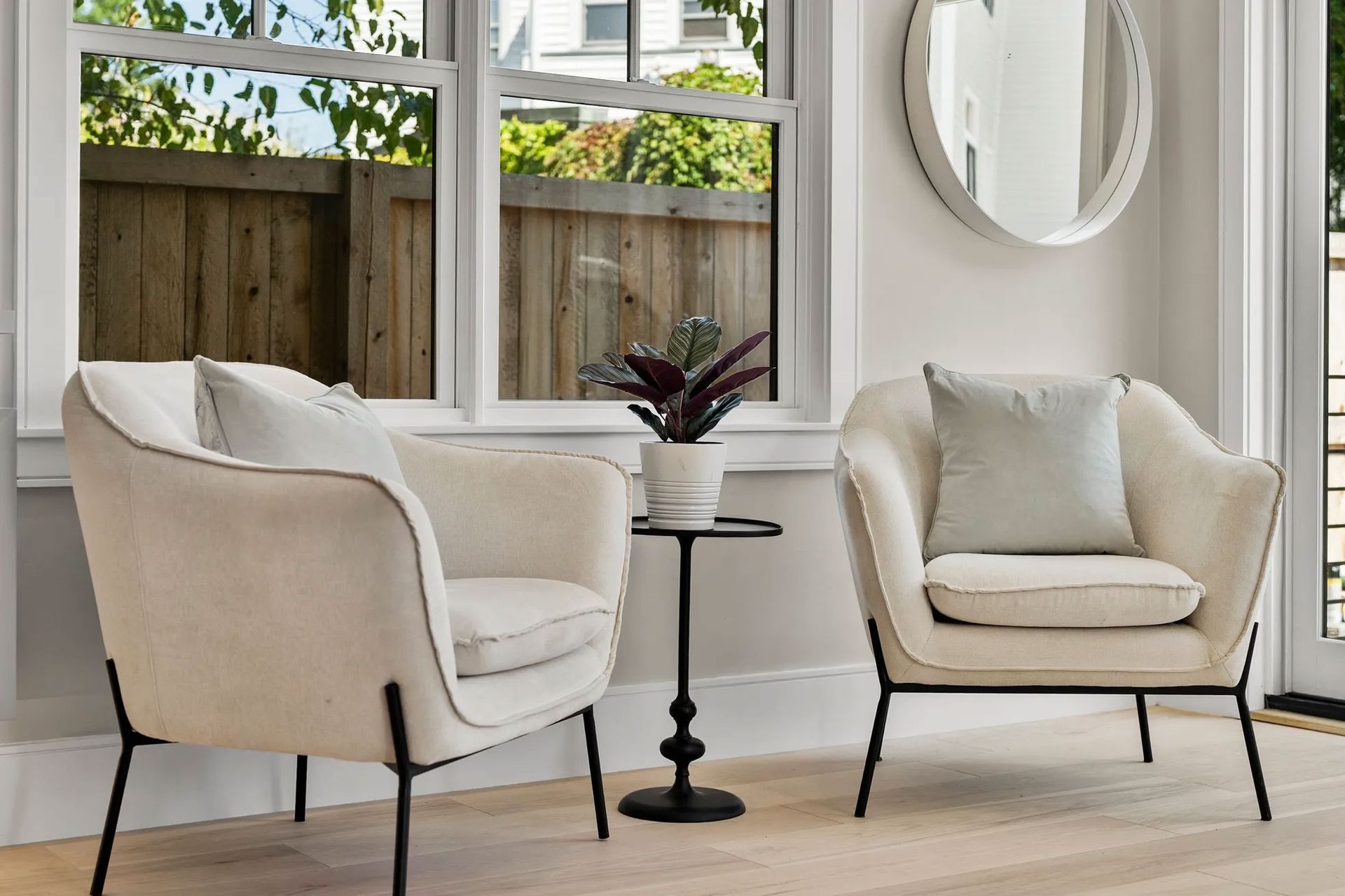 Two cream-colored armchairs with black legs, side table with a plant, window, and round mirror.