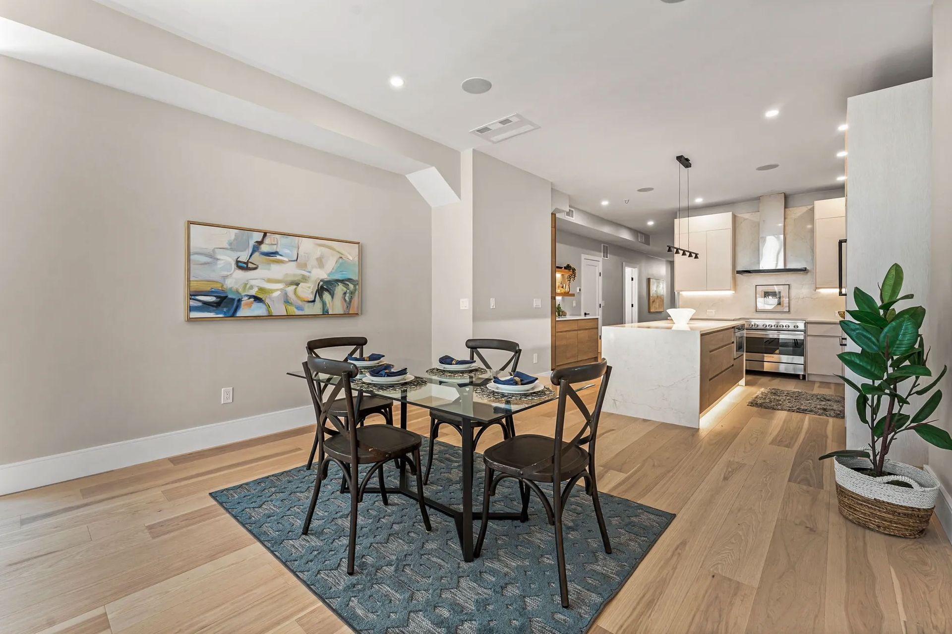 Dining room with table, chairs, rug, art, and open kitchen. Light wood floor and neutral walls.