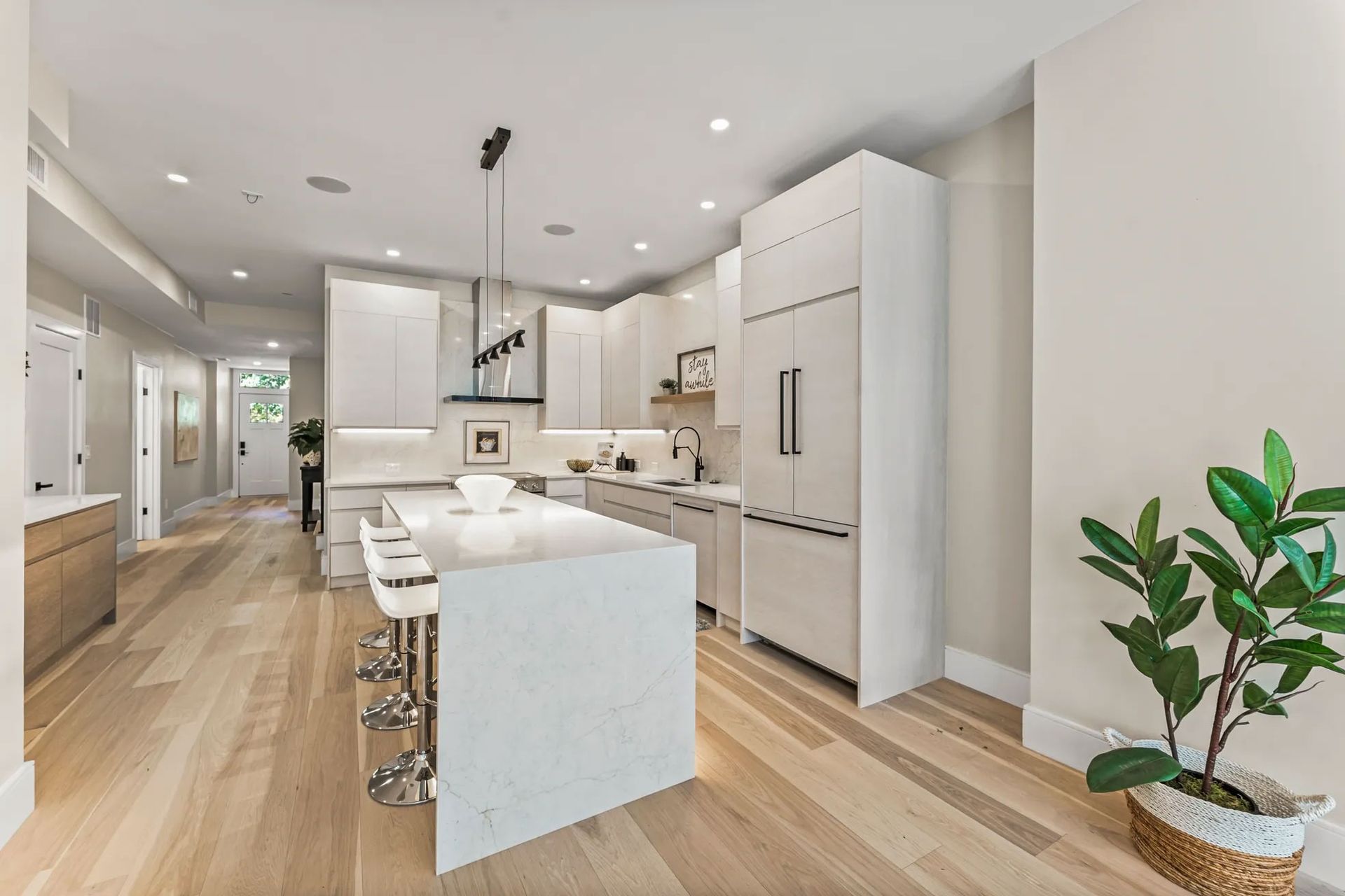 Modern white kitchen with island, stainless steel appliances, and wood flooring. A plant sits in the foreground.