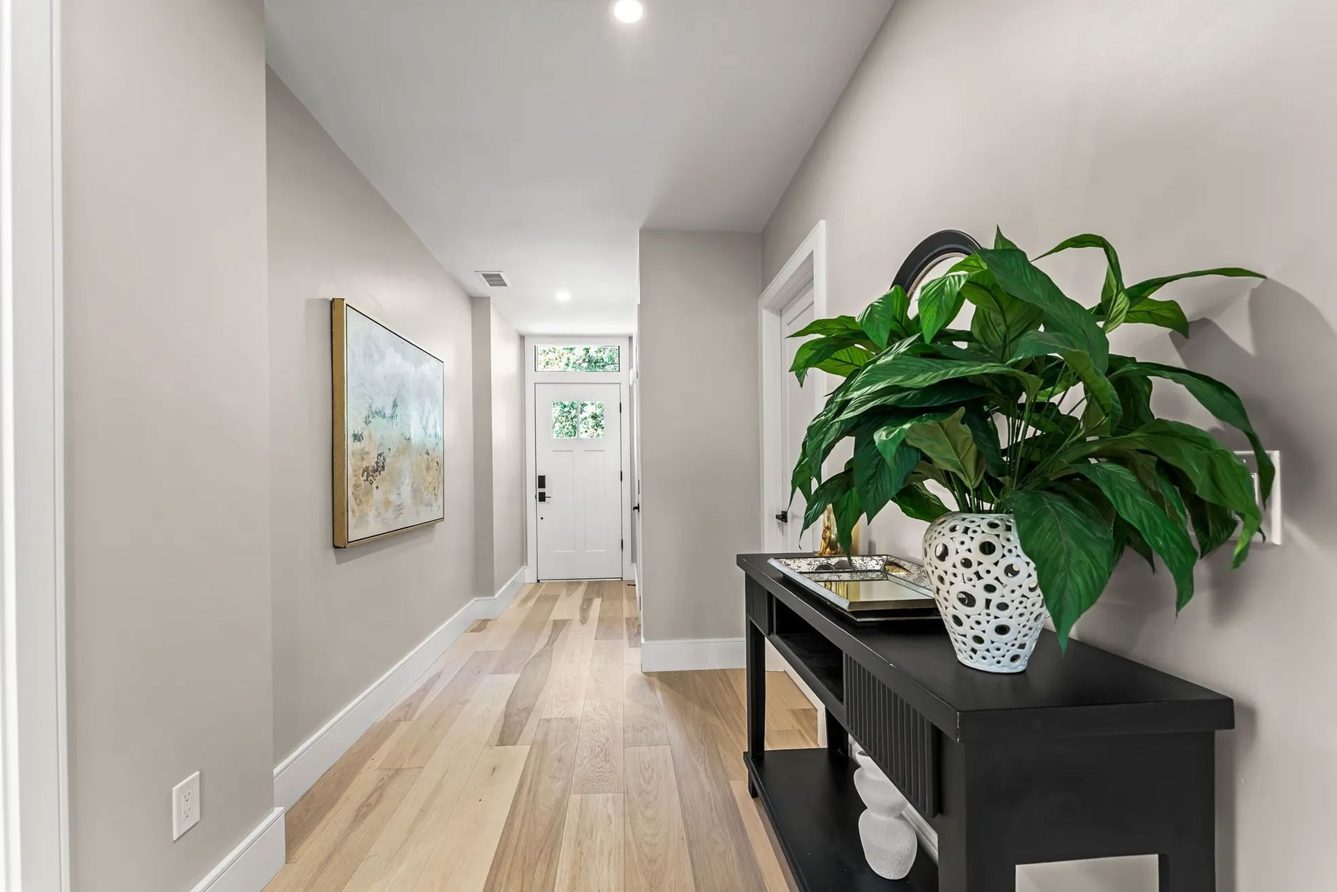 Hallway with light wood floors, neutral walls, and a black console table with a large potted plant.